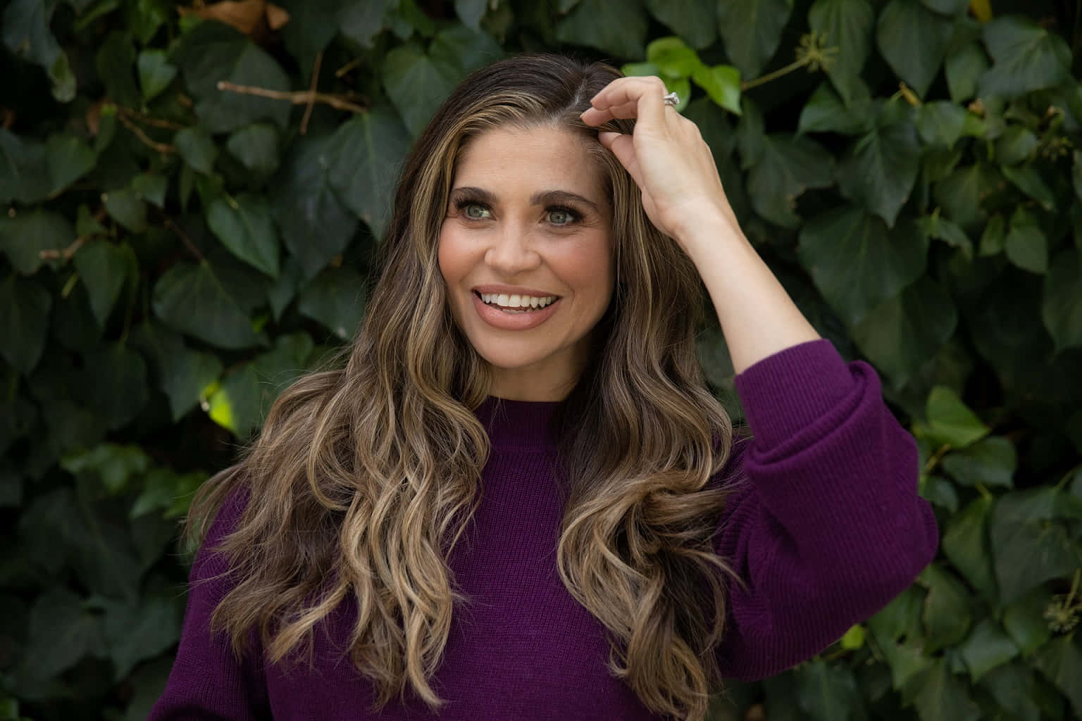 Danielle Fishel 桌布 壁紙 — 一位微笑的長波浪髮女性穿著紫色毛衣，輕輕地將頭髮觸碰在茂密的綠色常春藤牆上。