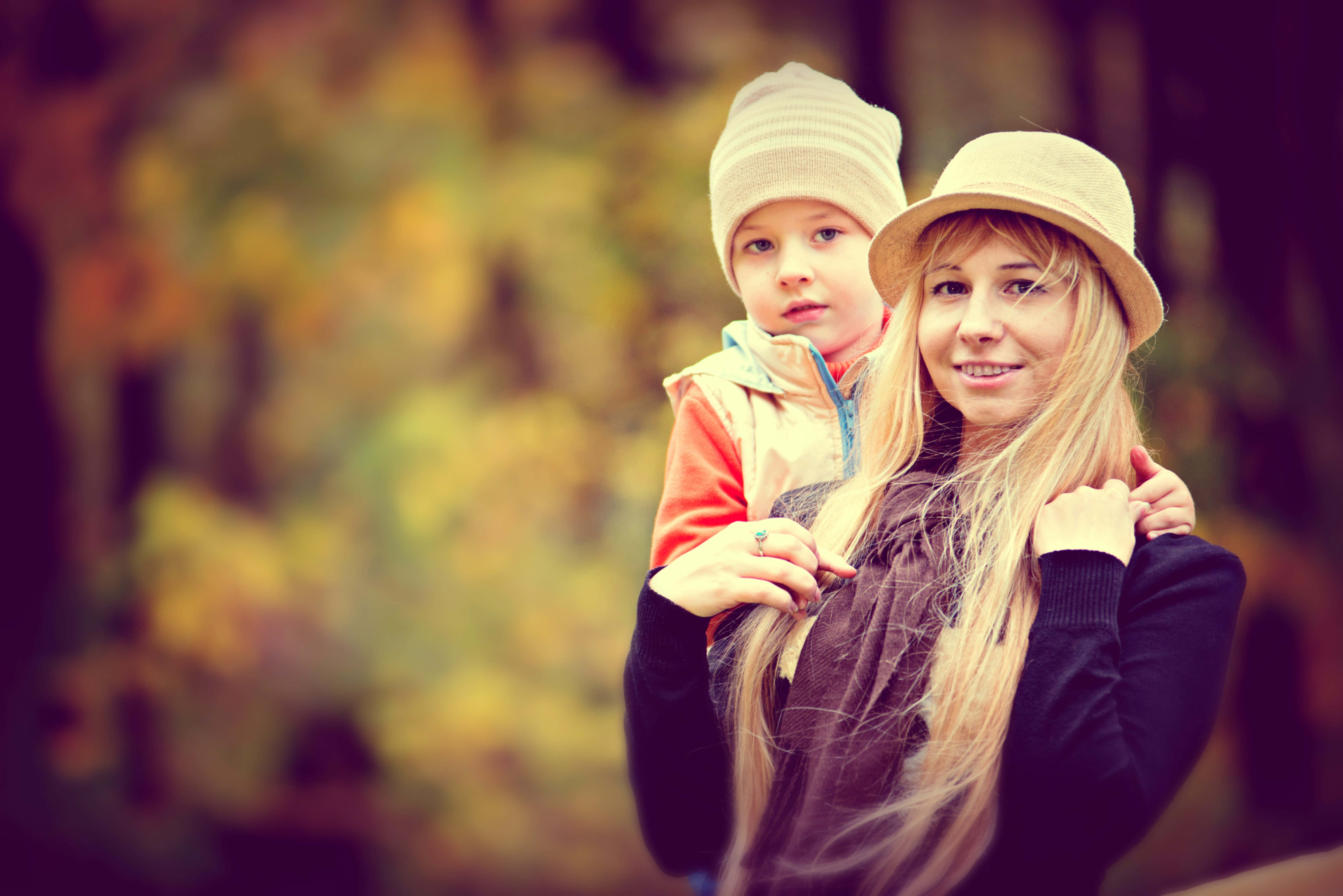 Mom 桌布 壁紙 — 一位微笑的長金髮女性，在柔和模糊的秋日森林背景下，將一個孩子扛在肩上。