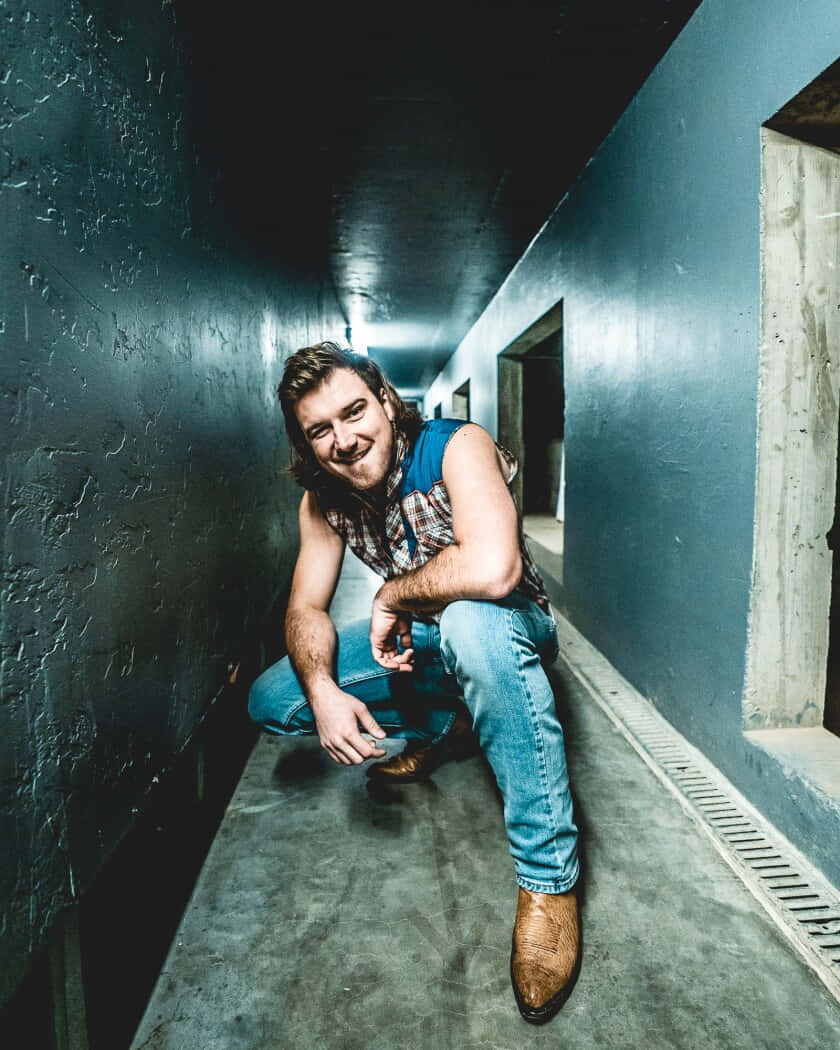 Morgan Wallen 桌布 壁紙 — 一位微笑的長髮男子蹲在狹窄的工業走廊中，牆壁有紋理，地板是混凝土。