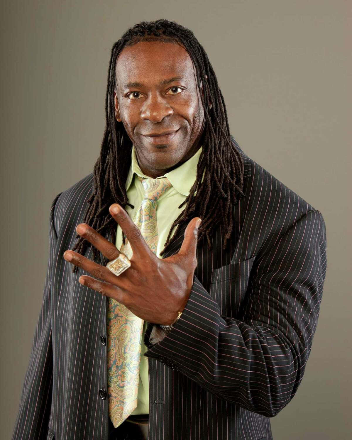 American Professional Wrestler Booker T Portrait 壁紙 — 一位微笑的長髮辮男子身穿細條紋西裝和圖案領帶，用戴著大戒指的手做手勢。