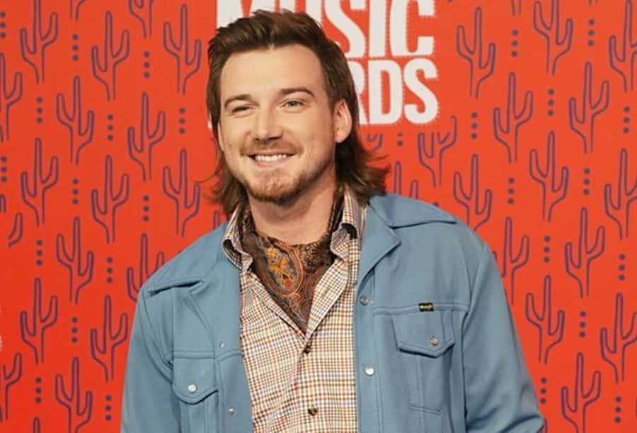Morgan Wallen 桌布 壁紙 — 一位微笑的長髮鬍鬚男，身穿牛仔外套內搭格仔恤衫，背景係鮮紅色仙人掌圖案。