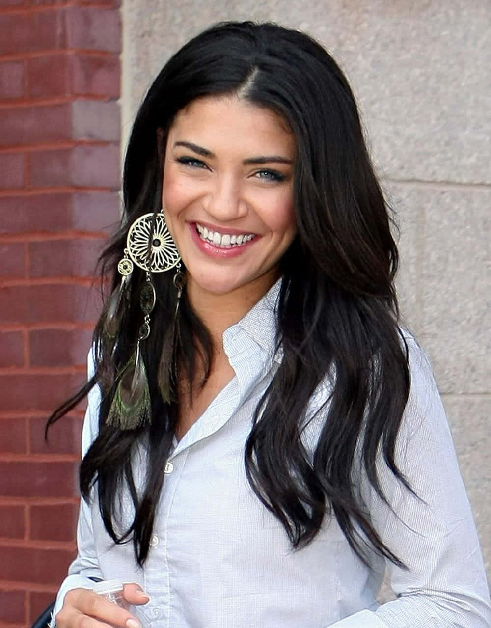 jessica szohr 桌布 壁紙 — 一位微笑的長黑髮女性，戴著大型華麗耳環，站在磚石牆前。