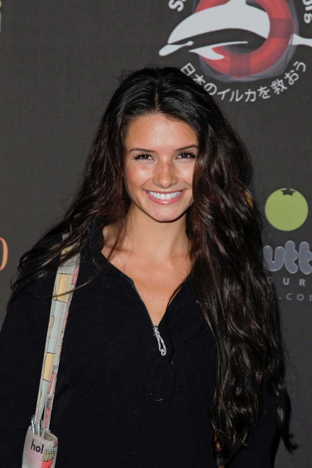 Alice Greczyn 桌布 壁紙 — 一位微笑的長黑髮女性在品牌背景的活動中穿著黑色拉鍊上衣。