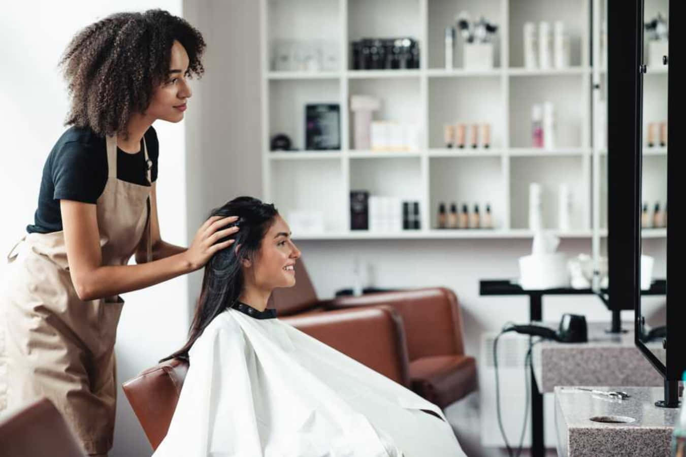 Hair Salon 桌布 壁紙 — 一位微笑的長黑髮客戶坐在沙龍椅上，一位捲髮造型師正在輕柔地按摩她的頭皮。