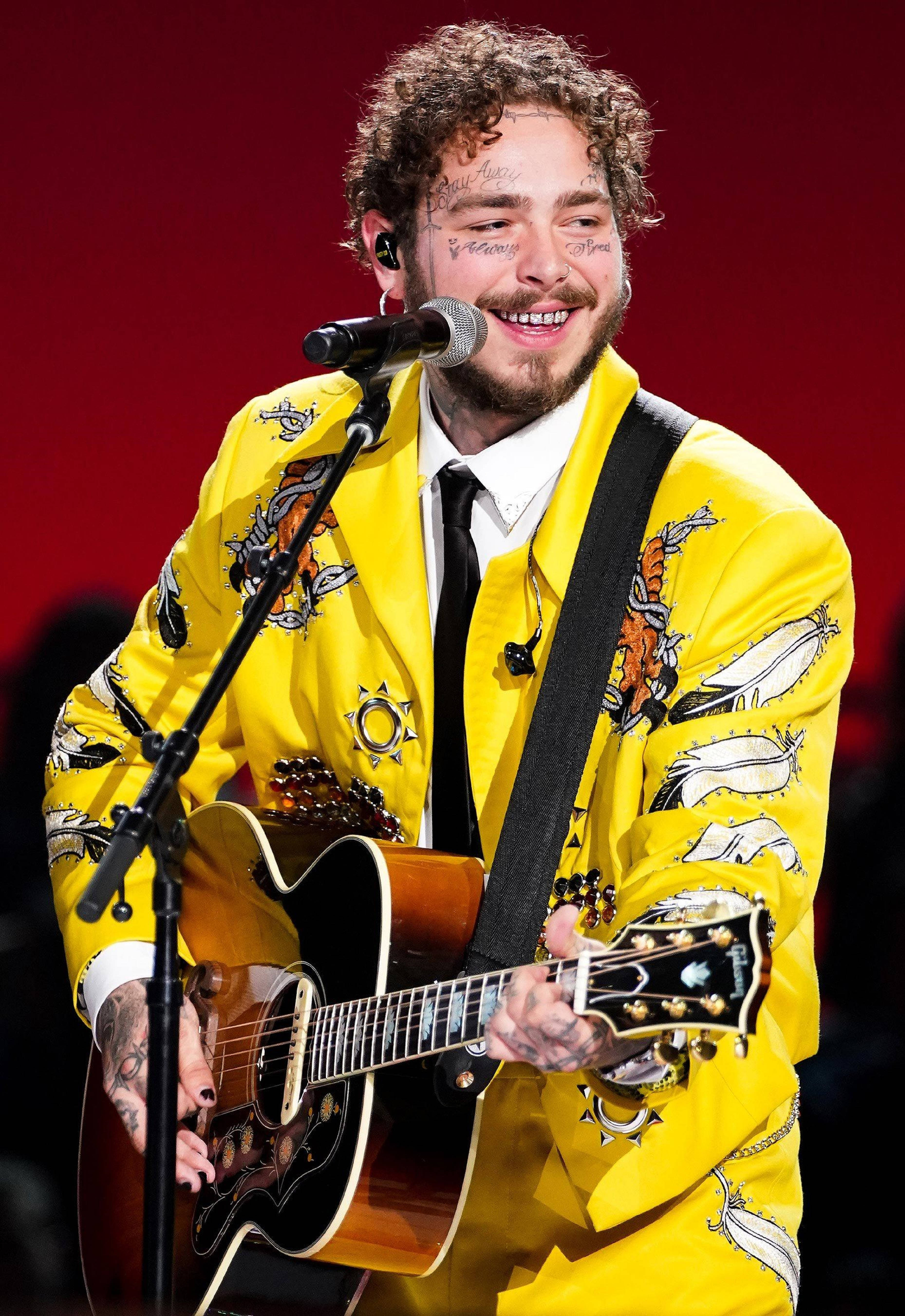 Post Malone Elvis Tribute 2019 壁紙 — 一位微笑的音樂家穿著亮黃色刺繡外套，在紅色燈光的舞台上彈奏原聲結他。