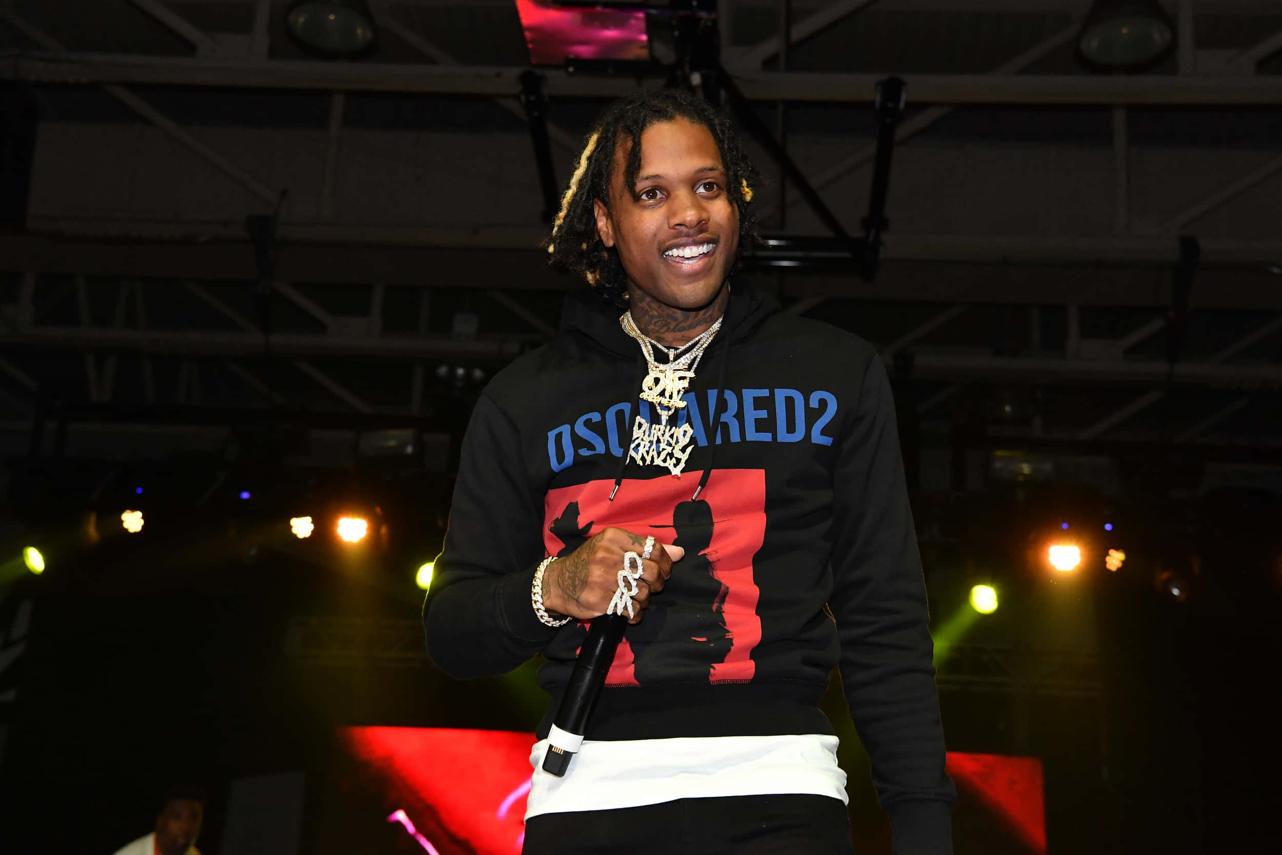 lil durk 桌布 壁紙 — 一位微笑的饒舌歌手穿著黑色連帽衫，在燈光昏暗的舞台上表演，頭頂上有彩色聚光燈和可見的桁架結構。