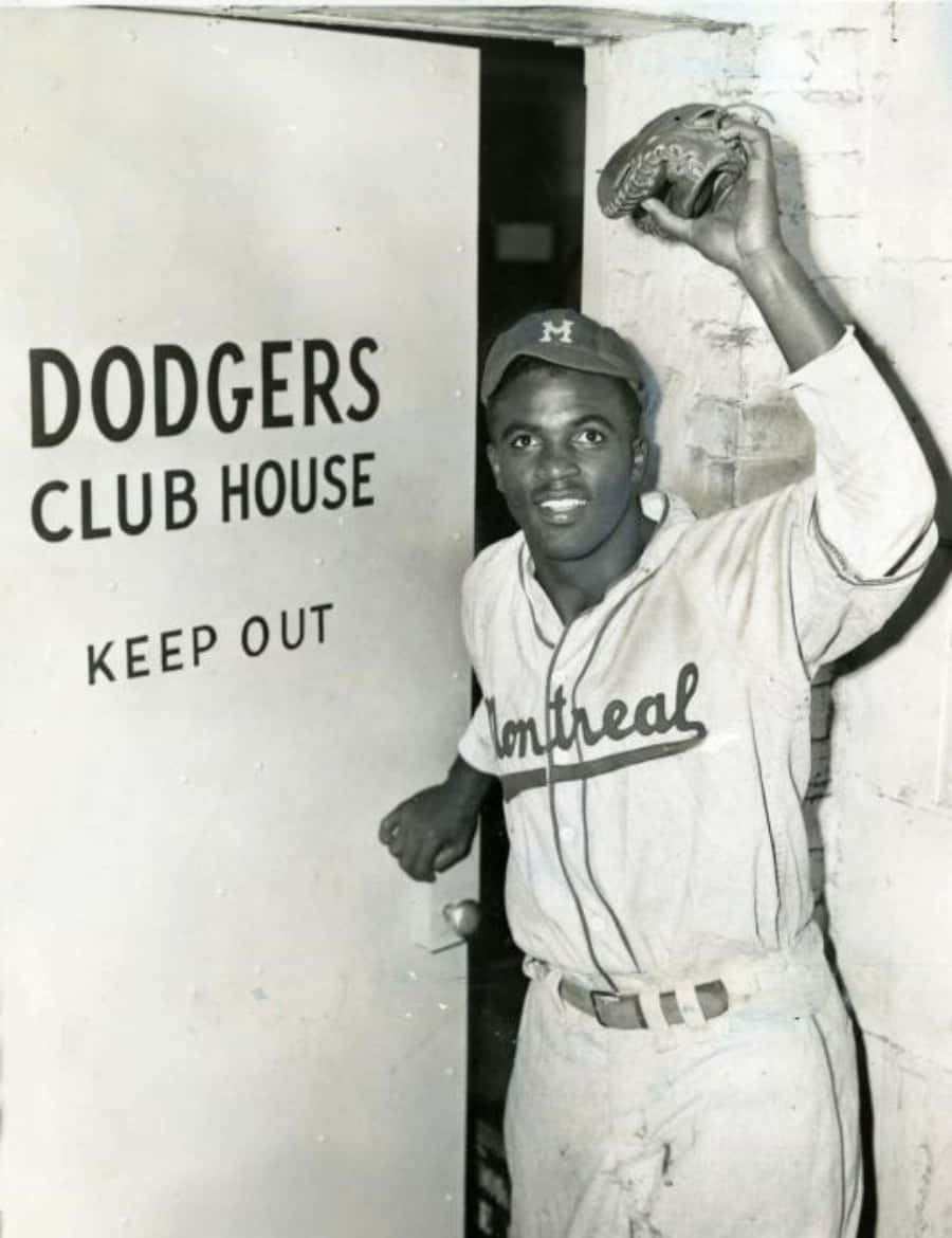 Jackie Robinson 桌布 壁紙 — 一位微笑的黑人棒球運動員穿著蒙特利爾隊服，在寫有 'DODGERS CLUB HOUSE KEEP OUT' 的門旁舉起手套。