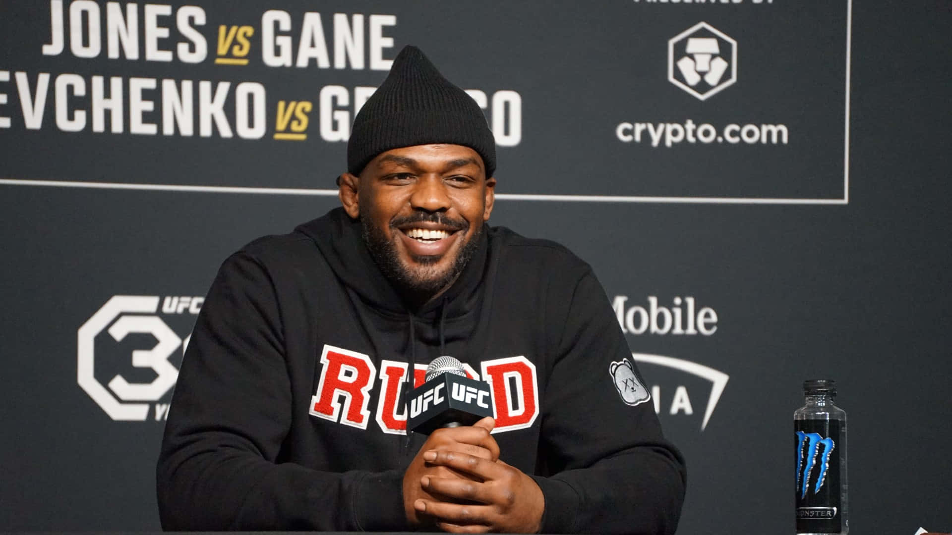 UFC 桌布 壁紙 — 一位微笑的黑人男子戴着黑色无檐便帽和连帽衫，在赞助商标志后面的新闻活动中对着UFC麦克风讲话。