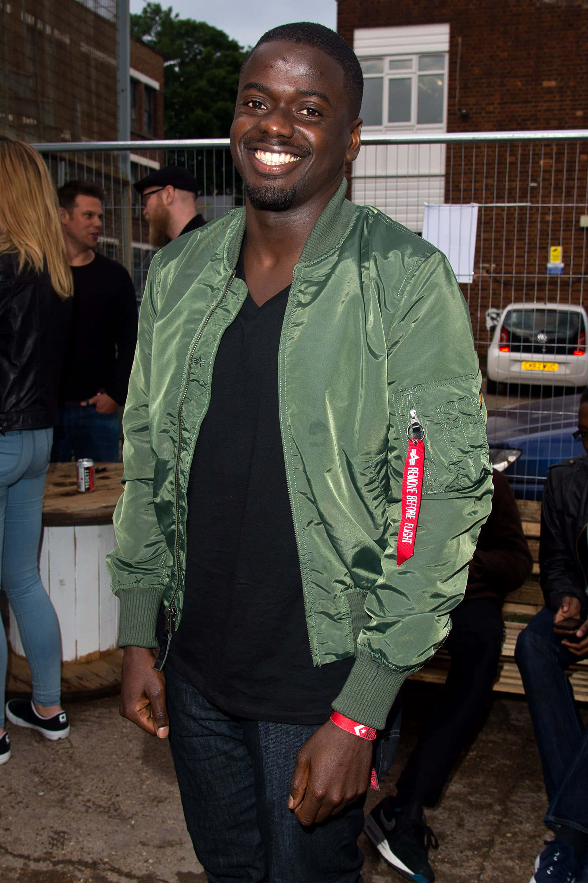 Daniel Kaluuya 桌布 壁紙 — 一位微笑的黑人男性穿著綠色飛行員夾克，在戶外活動中站立，背景是磚砌建築和圍欄。