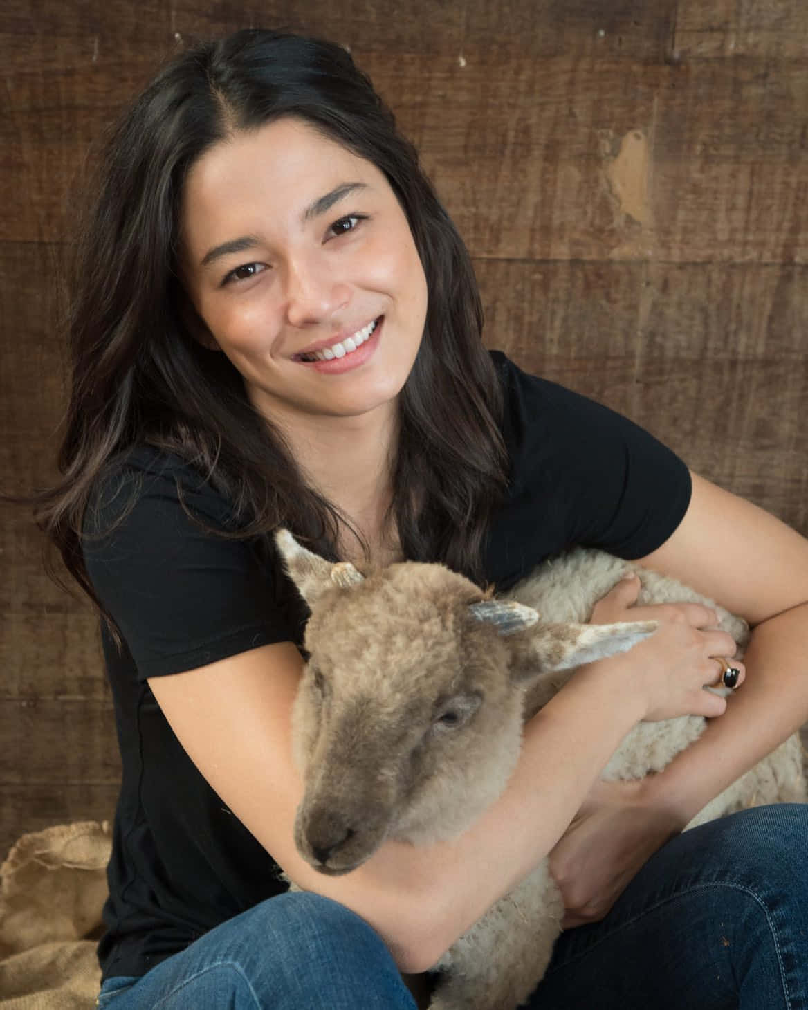jessica gomes 桌布 壁紙 — 一位微笑的黑衫女士在質樸的木質背景前輕輕懷抱一隻毛茸茸的小山羊。