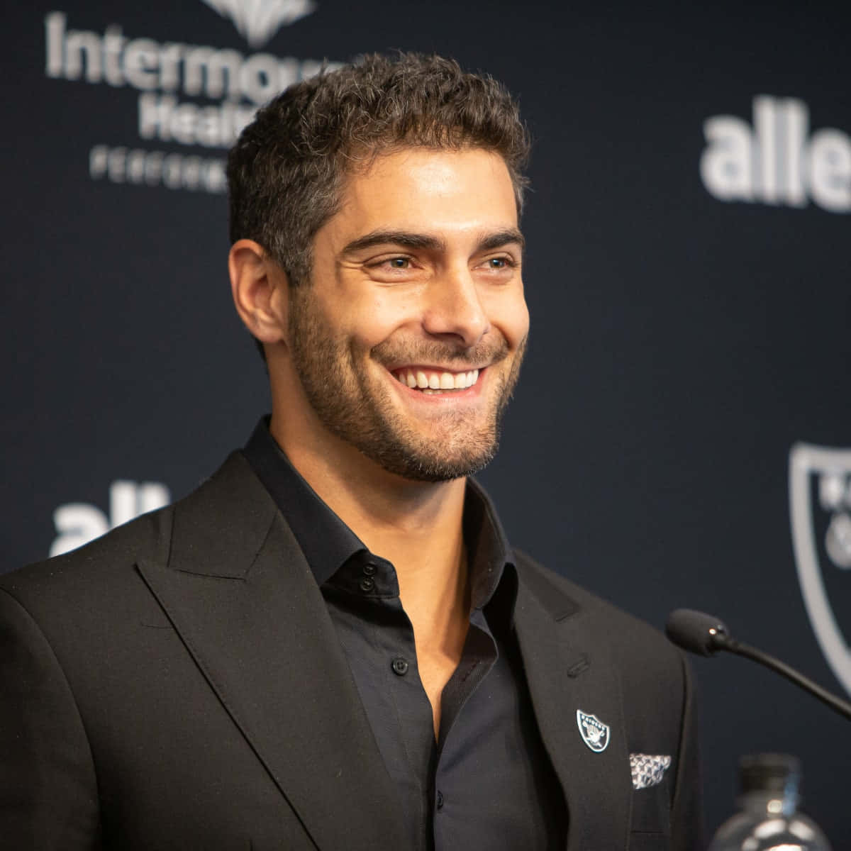 Jimmy Garoppolo 桌布 壁紙 — 一位微笑的黑西裝和襯衫男士站在咪高峰前，背景是深色品牌背景。