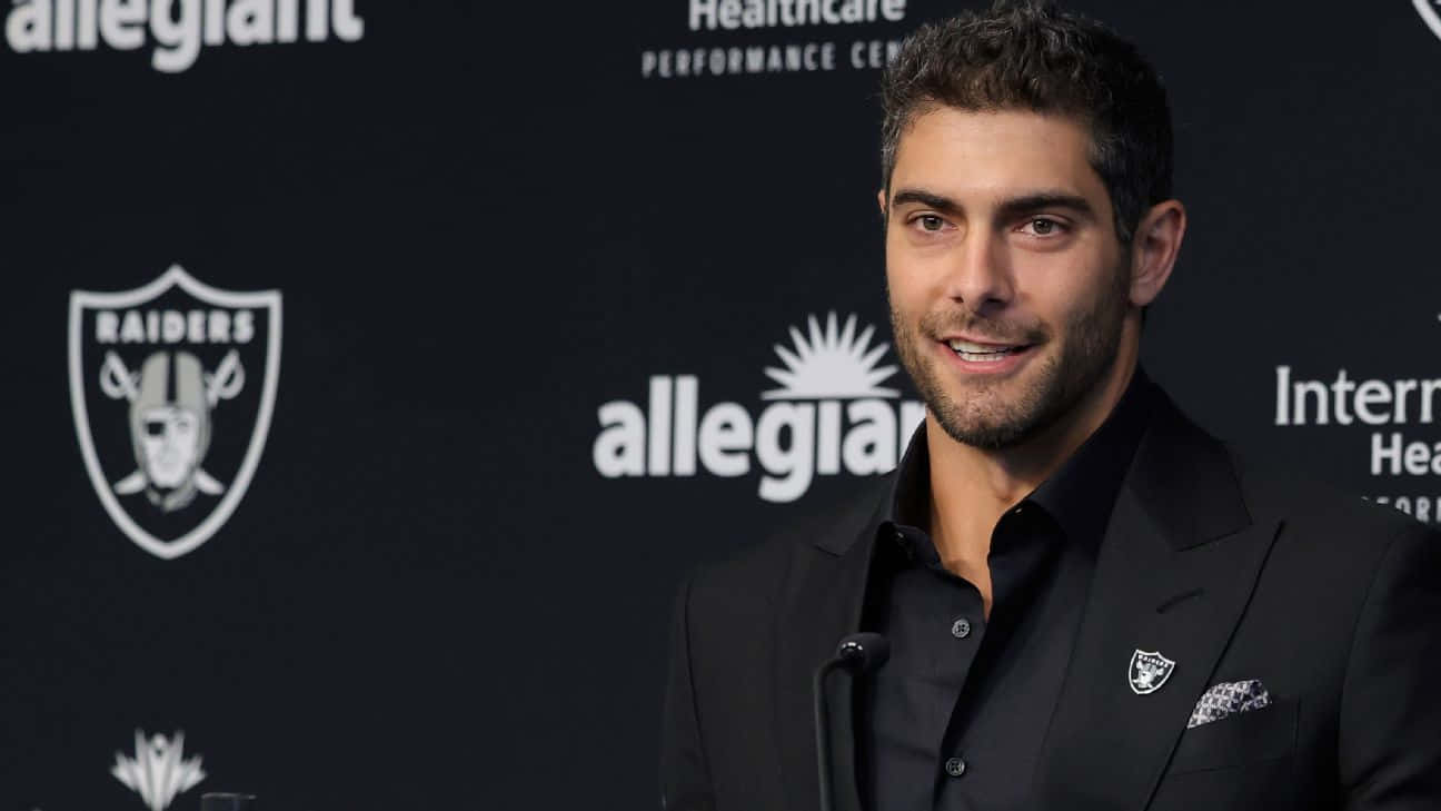 Jimmy Garoppolo 桌布 壁紙 — 一位微笑的黑西裝男士站在印有Raiders標誌和Allegiant品牌的背景前。