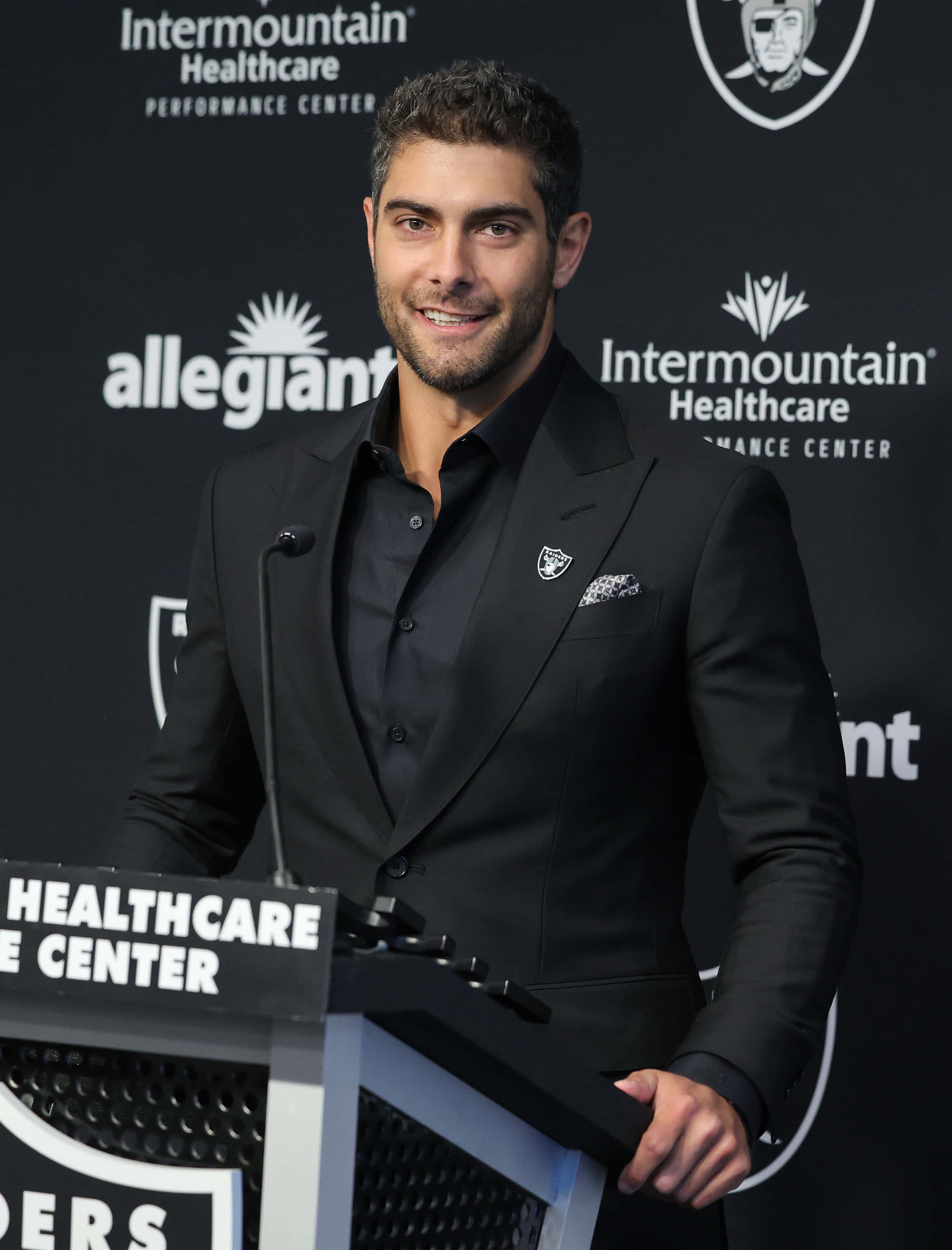 Jimmy Garoppolo 桌布 壁紙 — 一位微笑的黑西裝男士站在講台上，背景有Raiders標誌及品牌背景。