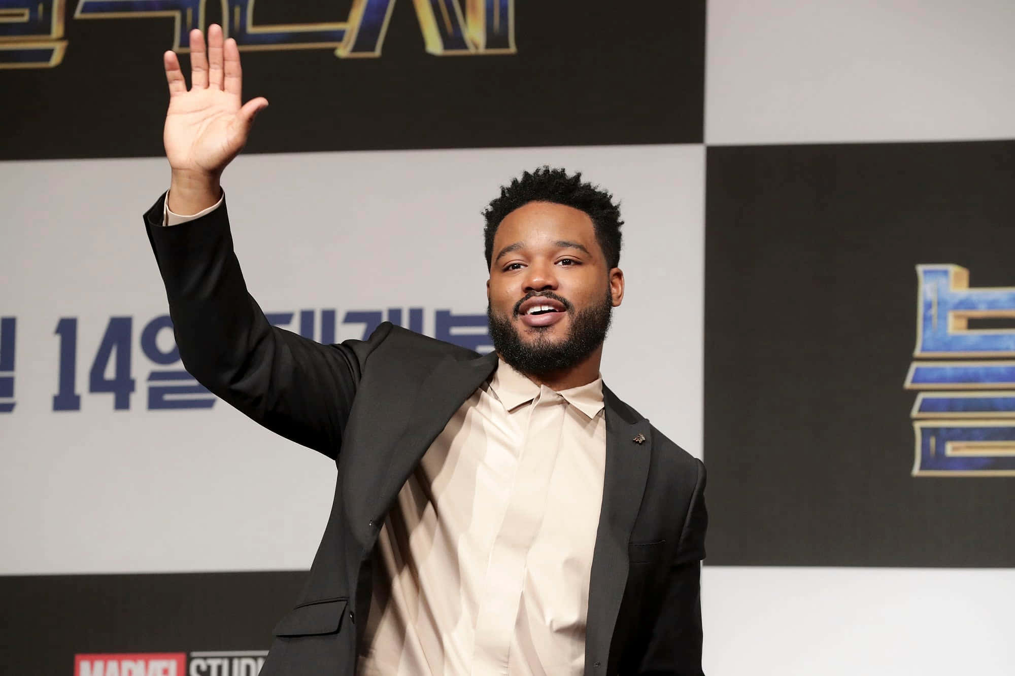 Ryan Coogler 桌布 壁紙 — 一位微笑的黑西裝男子在Marvel Studios活動中熱情揮手，背景為韓文文字。