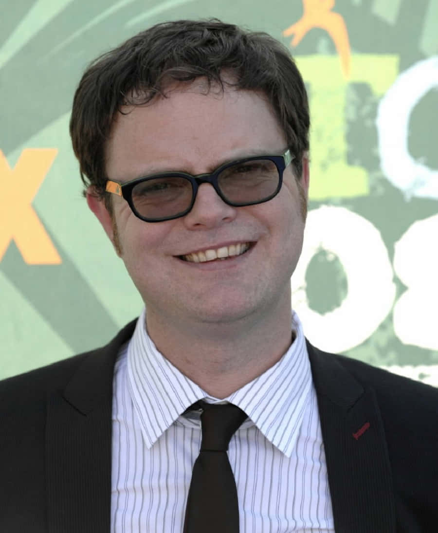 rainn wilson 桌布 壁紙 — 一位微笑的黑髮戴眼鏡男士，身穿黑色西裝和條紋襯衫，背景是色彩繽紛的活動場景。