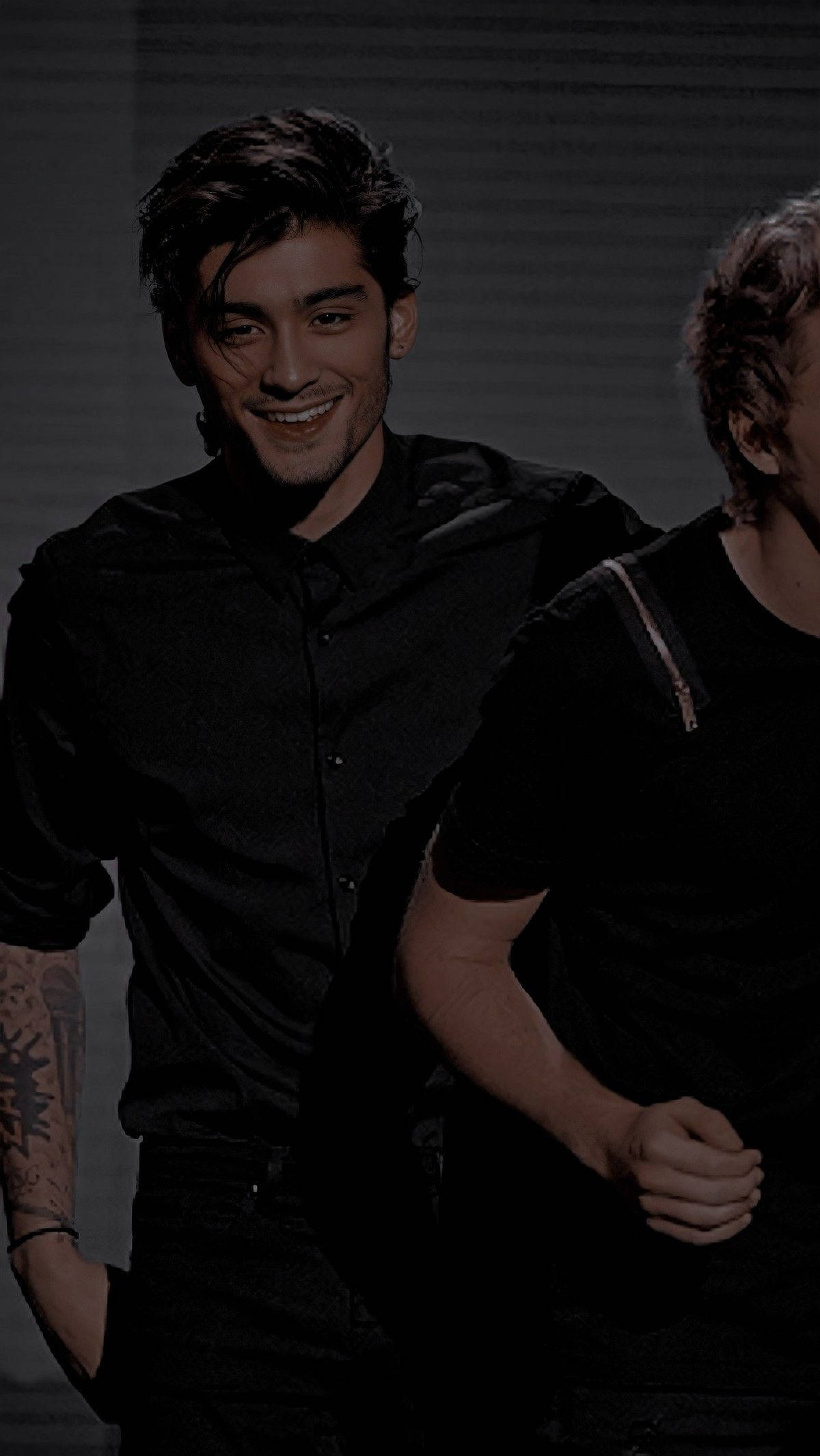 Zayn Malik 桌布 壁紙 — 一位微笑的黑髮男子，手臂上有紋身，穿著黑色襯衫，背景昏暗。