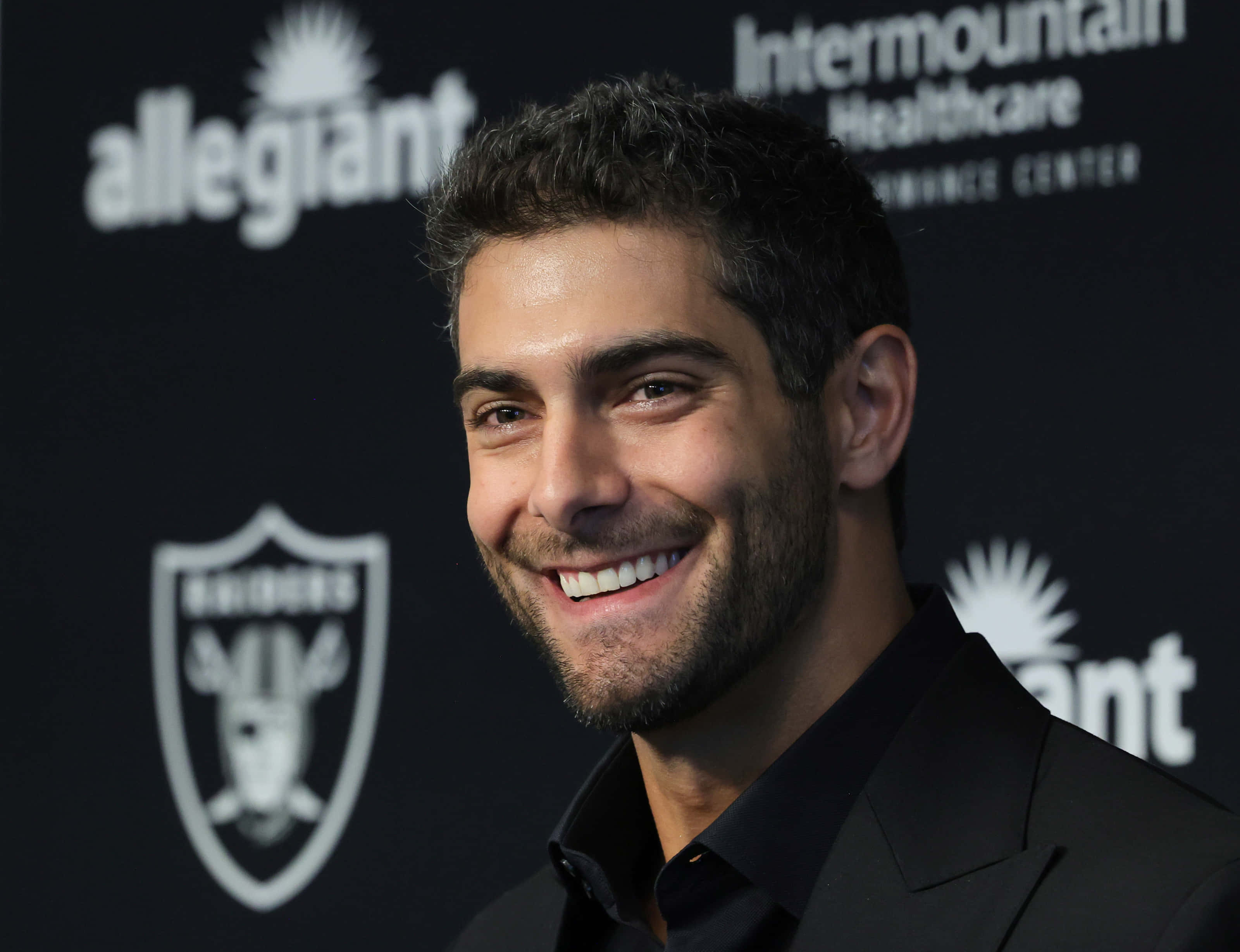 Jimmy Garoppolo 桌布 壁紙 — 一位微笑的黑髮鬍鬚男士身穿黑色西裝，站在黑色背景前，背景上有白色標誌，包括「Raiders」和「Intermountain Healthcare」。
