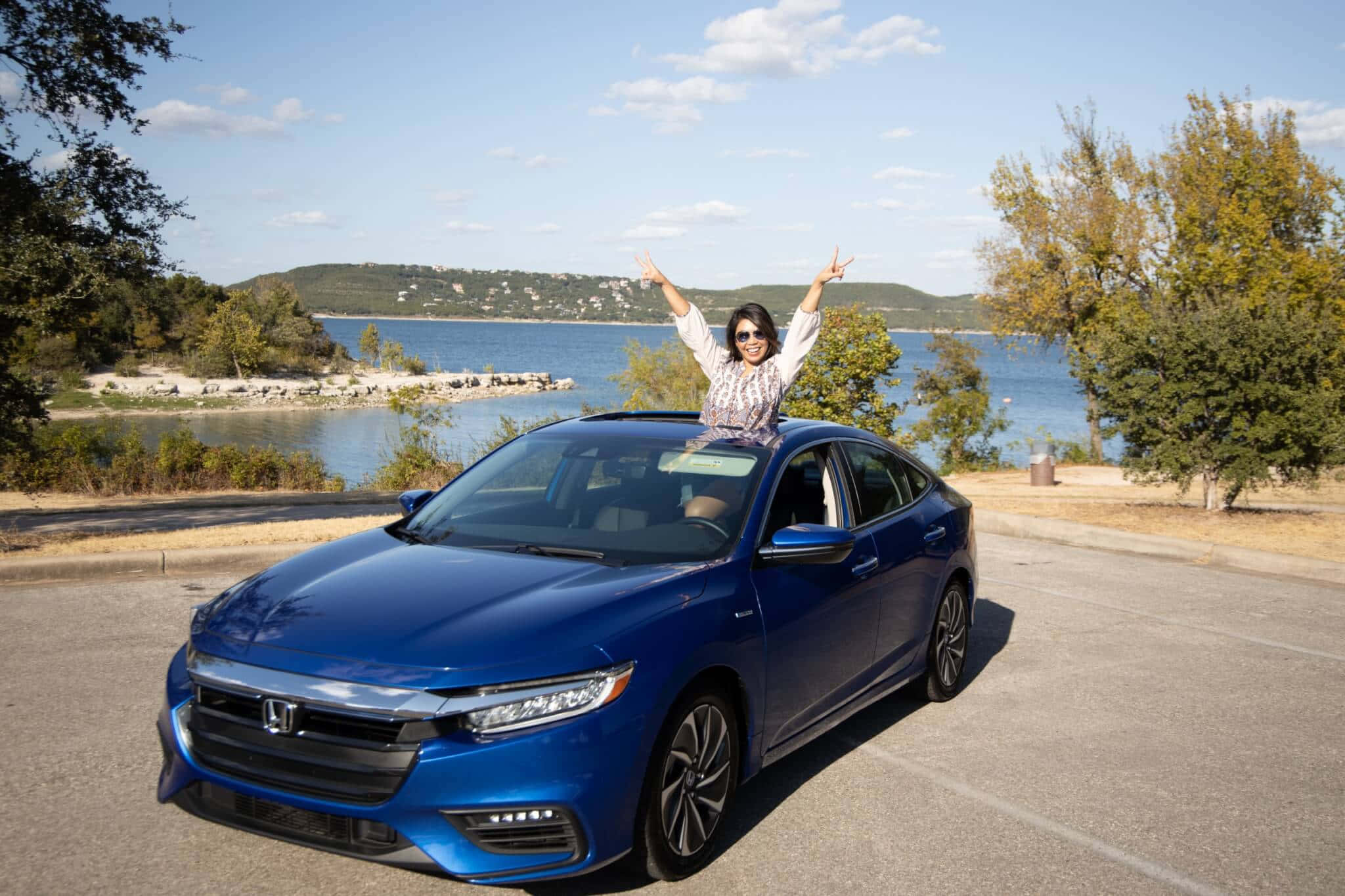 Honda Insight 桌布 壁紙 — 一位快樂的女性高舉雙臂，坐在一輛停在風景如畫的湖邊的藍色本田轎車上，天空湛藍。
