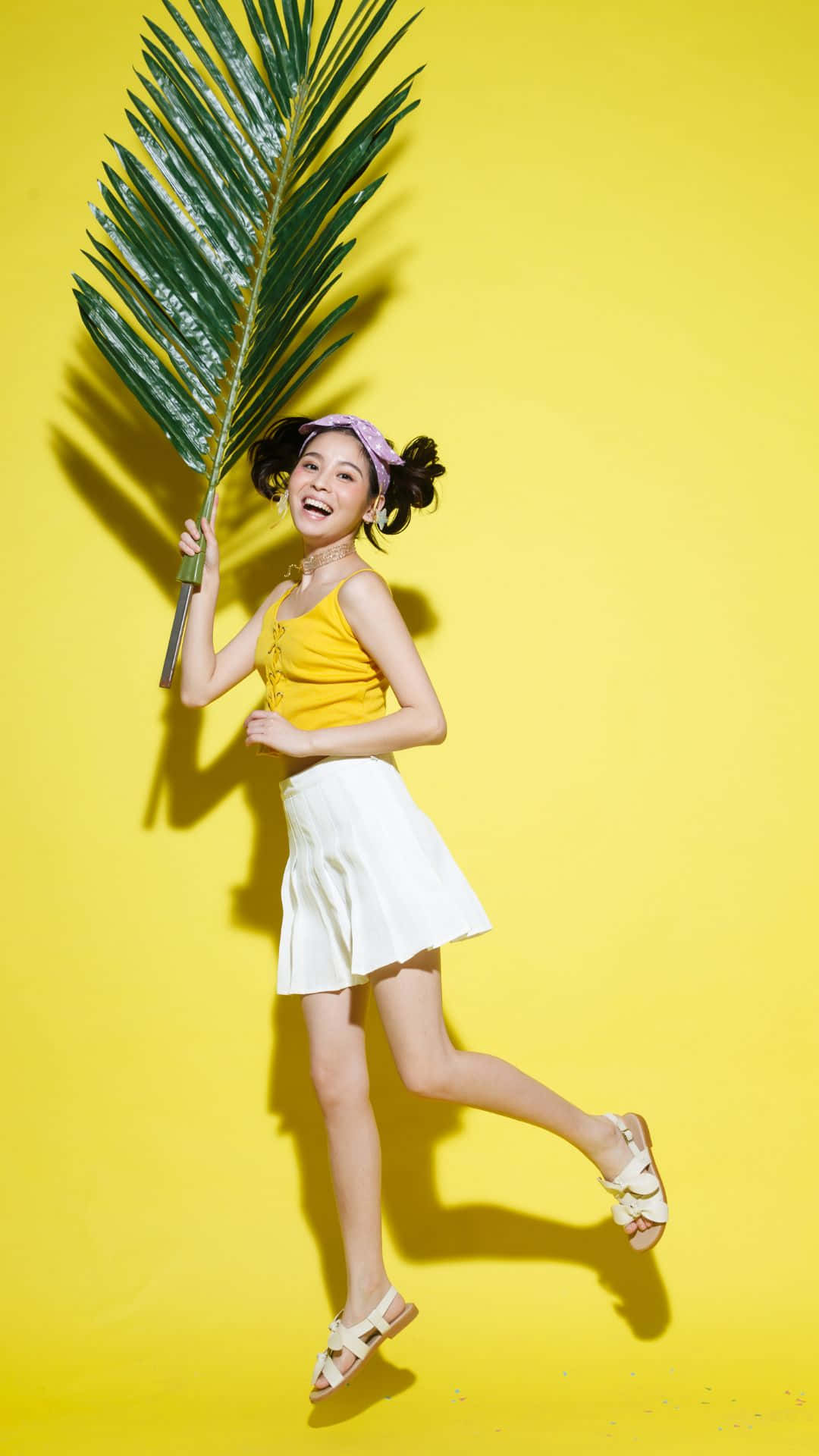 Happy iPhone 桌布 壁紙 — 一位快樂的年輕女性身穿黃色上衣和白色裙子，手持大片綠色棕櫚葉，在鮮黃色背景前跳躍。