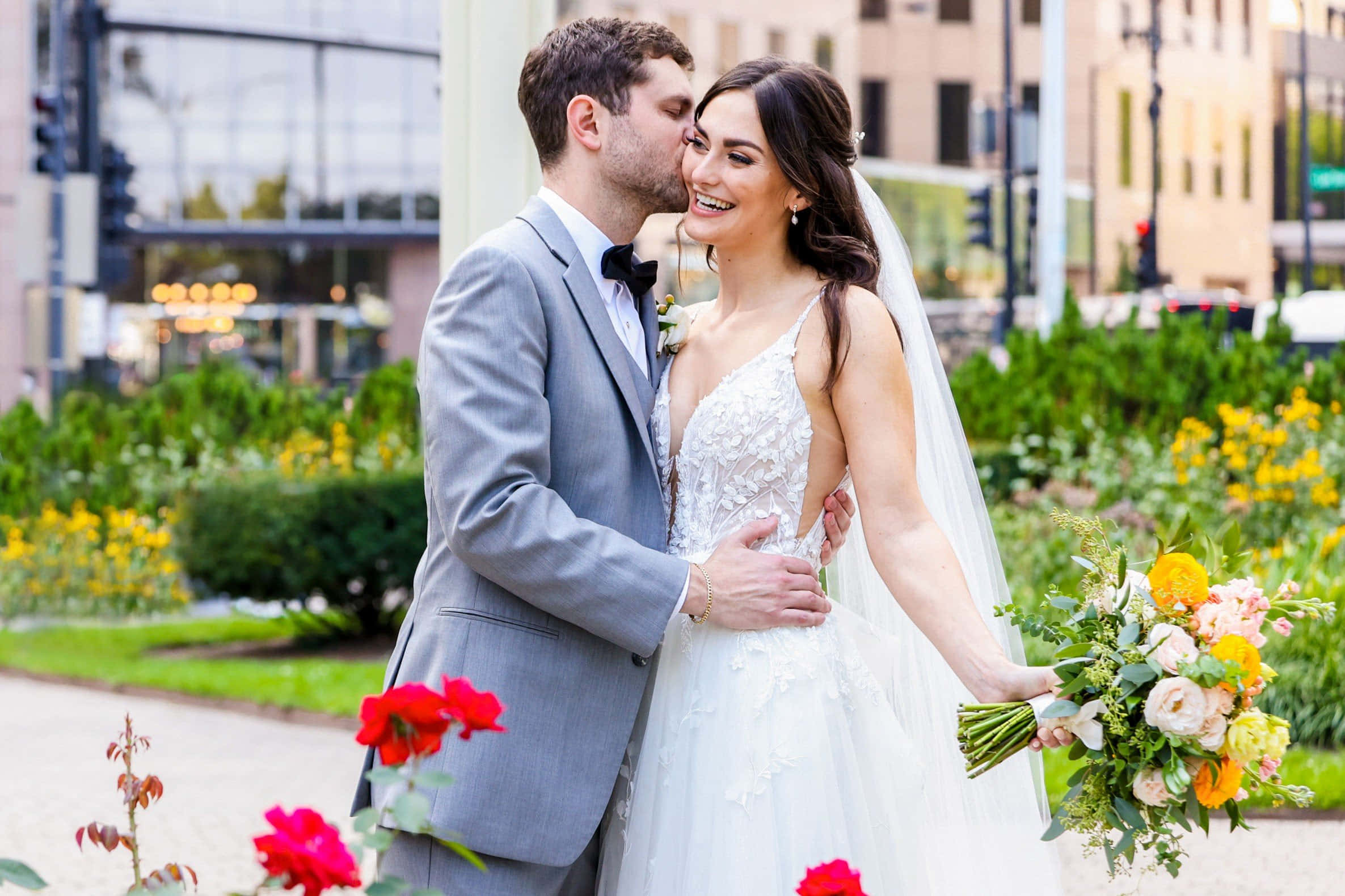Garden Wedding 桌布 壁紙 — 一位快樂的新娘在陽光普照的城市花園中，新郎親吻她的臉頰時大笑，背景有色彩繽紛的花朵和現代建築。