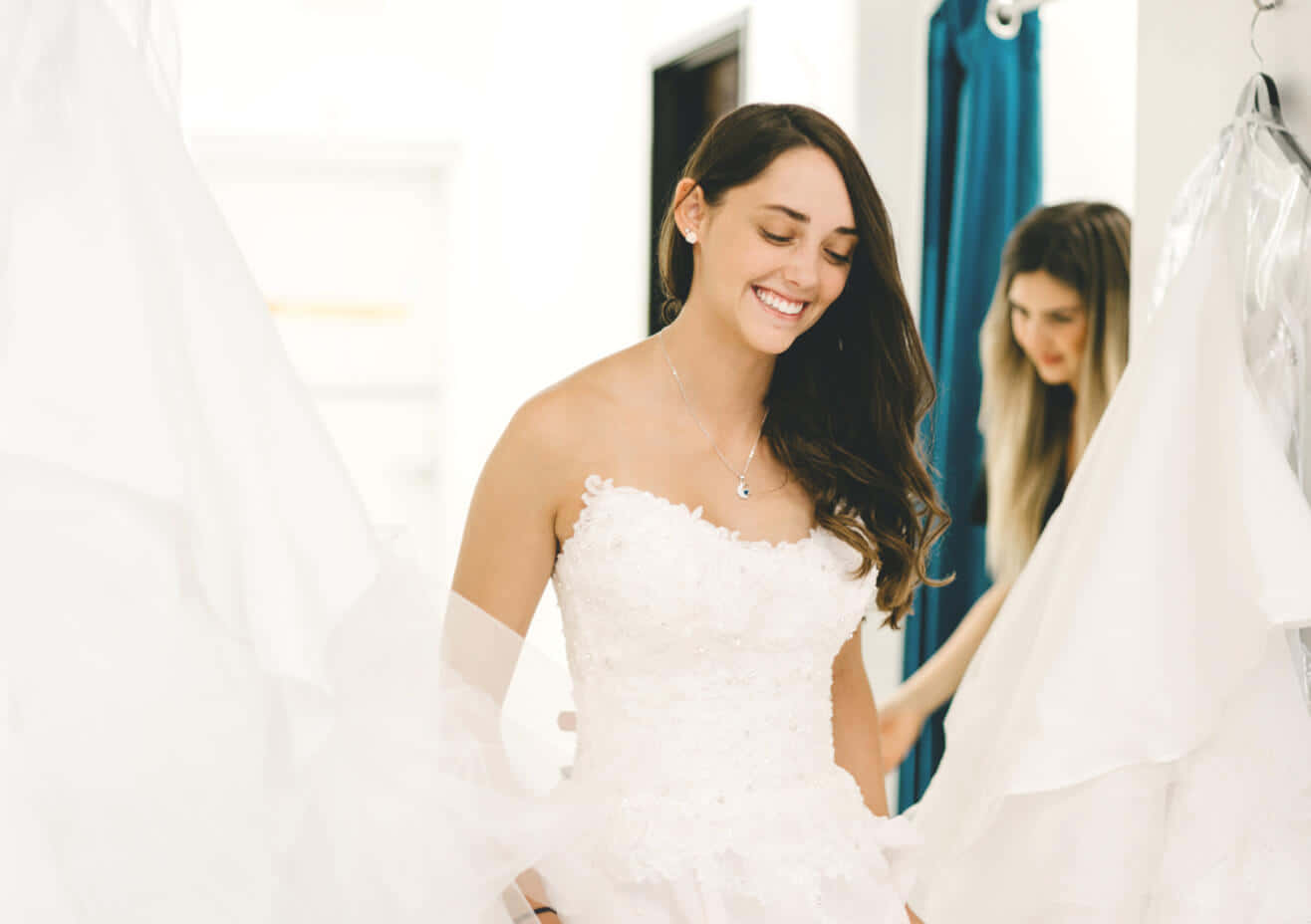 Bride's Dress 桌布 壁紙 — 一位快樂的新娘穿著白色蕾絲婚紗，在朋友附近試穿時微笑。