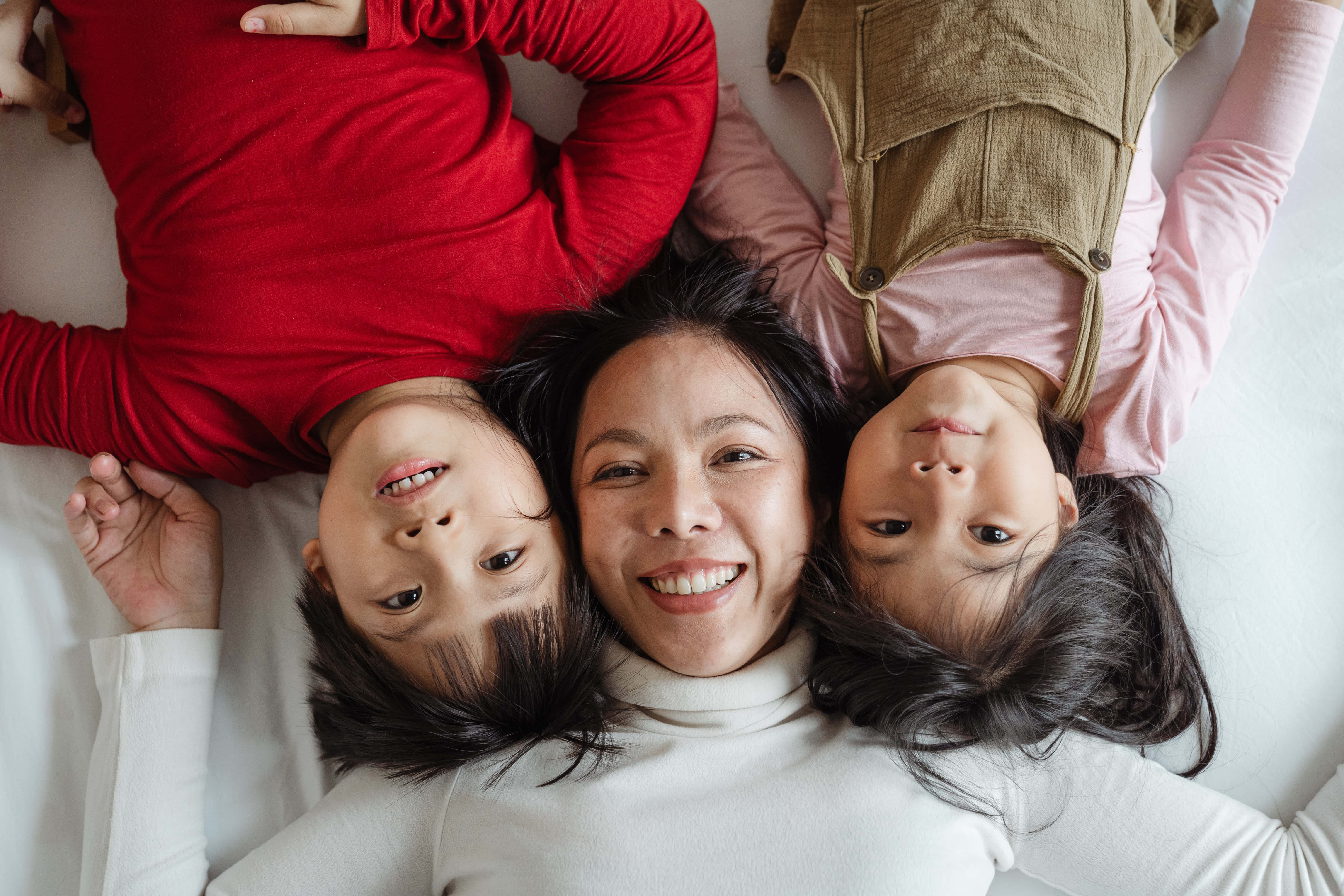 Mom 桌布 壁紙 — 一位快樂的母親和她的兩個孩子一起躺在白色床上，微笑著看向鏡頭，是一張溫馨的俯拍照片。