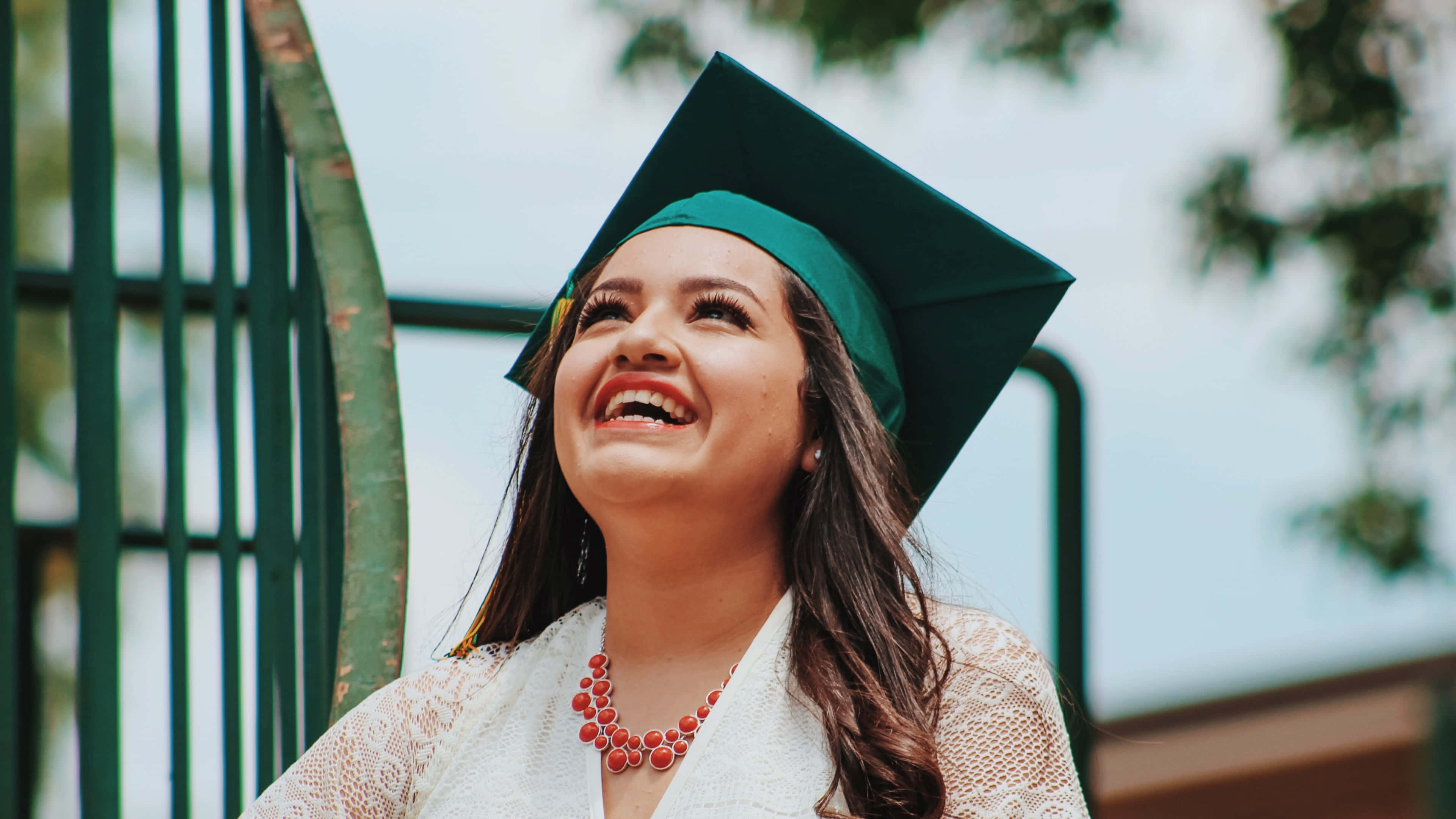 Graduation Ceremony 桌布 壁紙 — 一位快樂的畢業生戴着綠色四方帽和白色畢業袍，在戶外樹木和圍欄背景下開懷大笑。