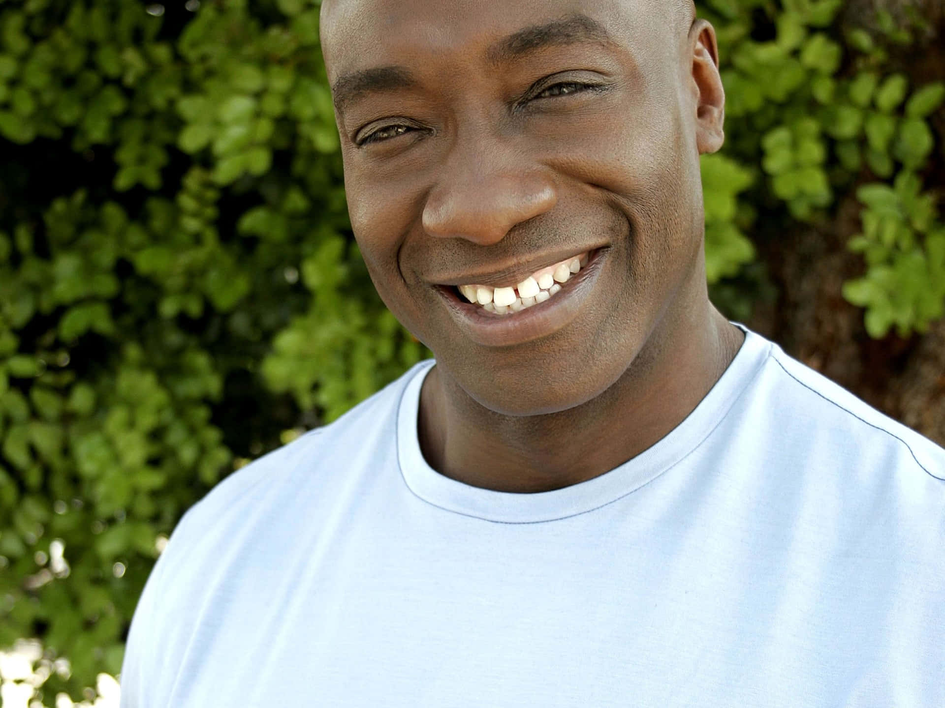 Michael Clarke Duncan 桌布 壁紙 — 一位快樂的黑人男性在茂密的綠色樹葉背景下露出燦爛的笑容。