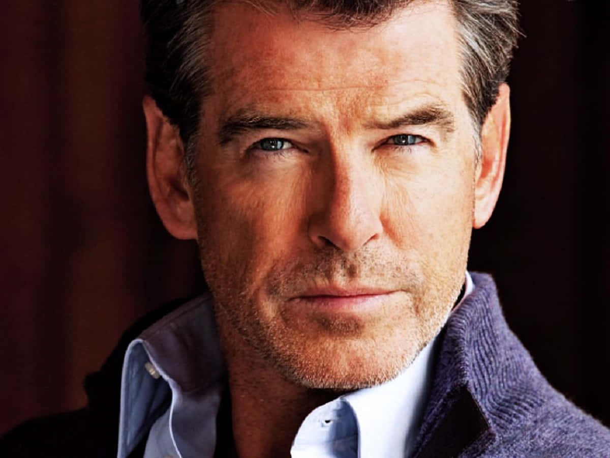 pierce brosnan 桌布 壁紙 — 一位成熟男士的 close-up 肖像，擁有強烈的藍色眼睛和鹽胡椒色頭髮，穿著紫色毛衣內搭淺藍色襯衫。