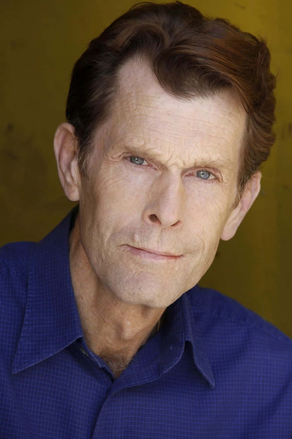 Kevin Conroy 桌布 壁紙 — 一位成熟男士的特寫肖像，擁有強烈的藍色眼睛和波浪形棕色頭髮，穿著藍色格仔襯衫，背景是溫暖的黃色。