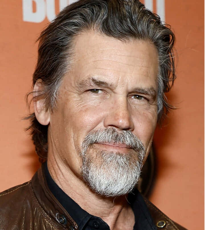 Josh Brolin 桌布 壁紙 — 一位成熟男士的特寫肖像，頭髮和鬍鬚花白，身穿棕色皮夾克，背景為橙色。