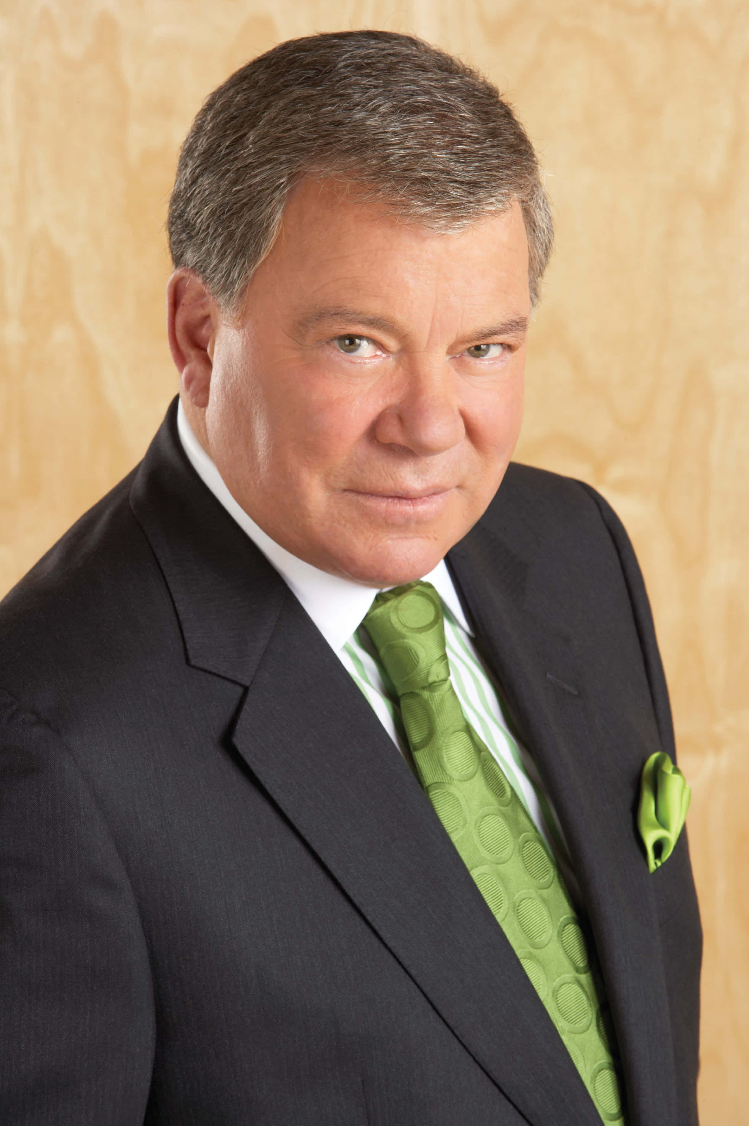 william shatner 桌布 壁紙 — 一位成熟男士身穿俐落黑色西裝和鮮綠色領帶，在溫暖的木鑲板背景前自信地擺姿勢。