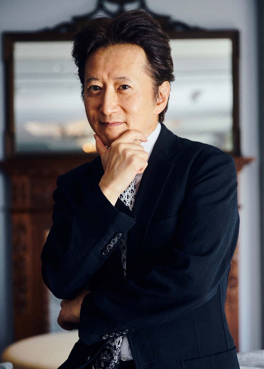 Hirohiko Araki 桌布 壁紙 — 一位成熟男士身穿深色西裝，在模糊的華麗室內背景下，手托下巴，擺出沉思的姿勢。