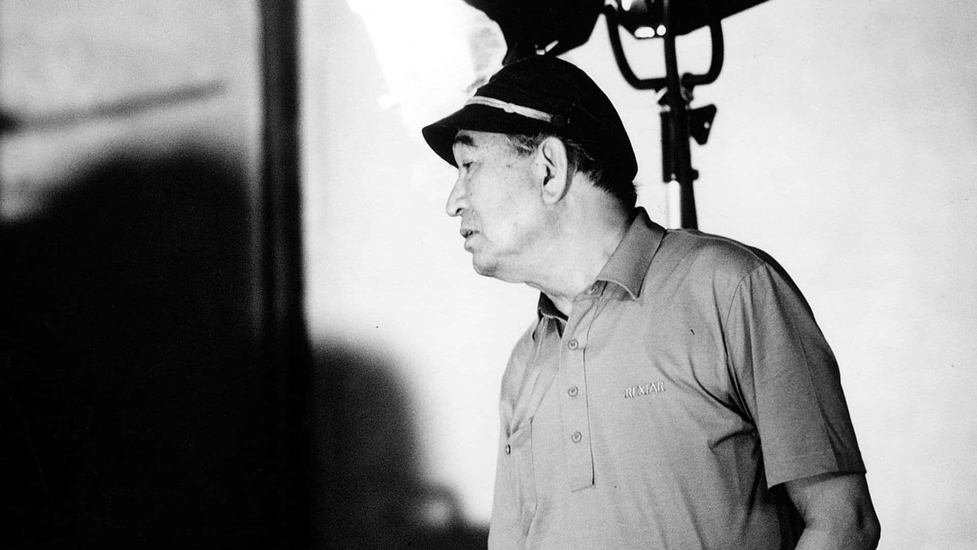 Akira Kurosawa 桌布 壁紙 — 一位戴帽子和Polo衫的老年男性站在攝影棚內，身後有燈光設備的黑白肖像。