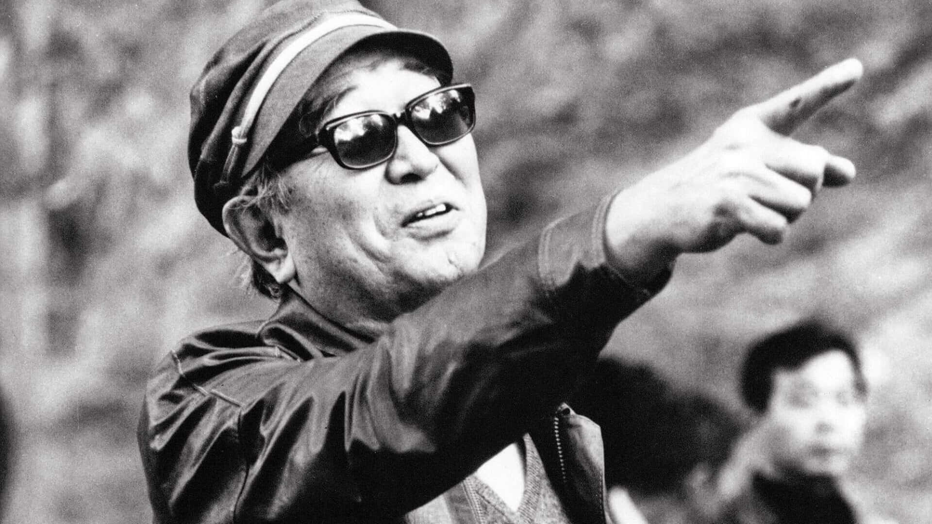 Akira Kurosawa 桌布 壁紙 — 一位戴帽子和太陽眼鏡的老年男子，強烈地指向前方的黑白照片。