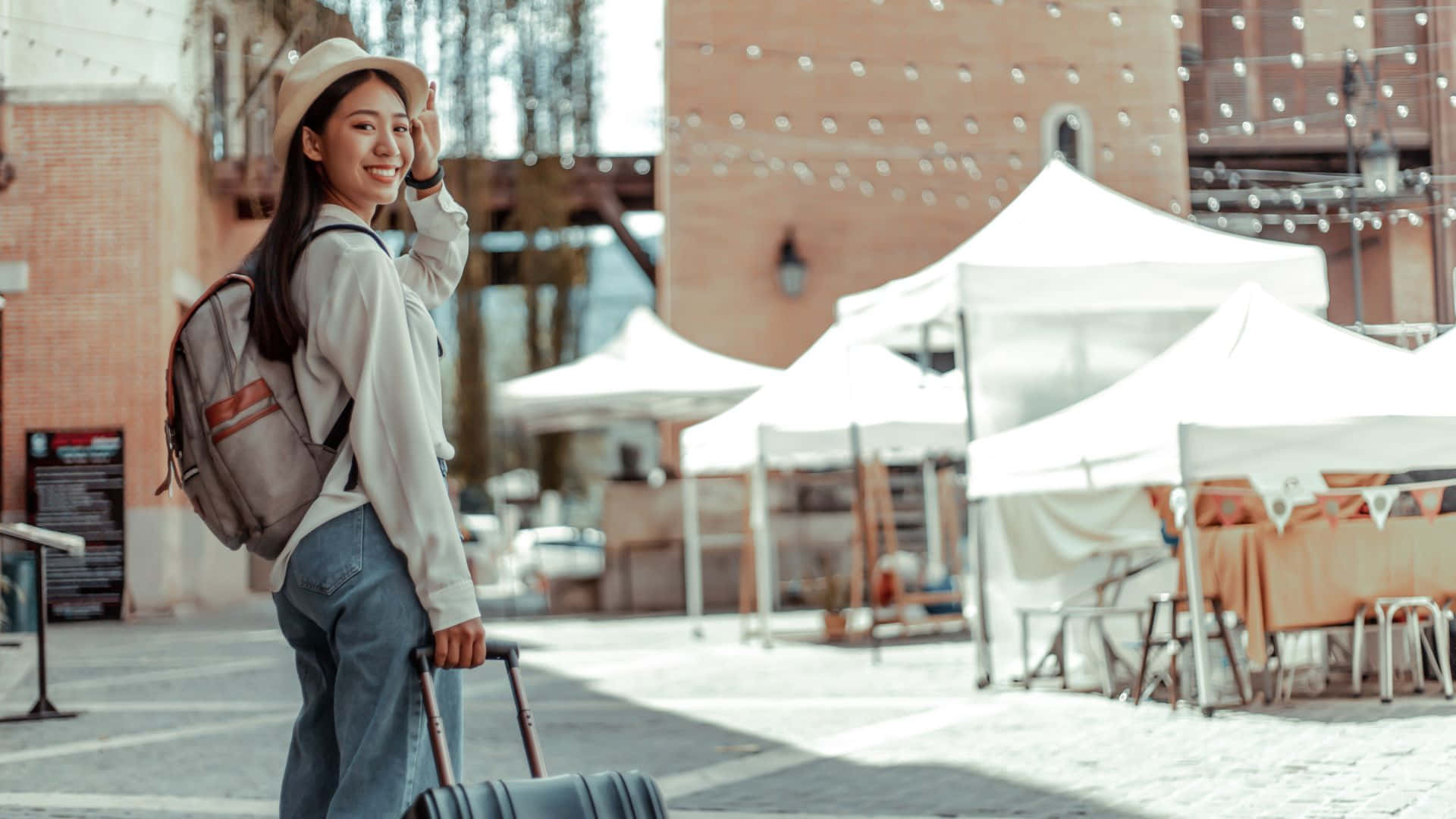 Beautiful Travel 桌布 壁紙 — 一位戴帽子和穿牛仔褲的年輕微笑女子，在陽光明媚的城市廣場上拉著行李箱，廣場上有白色市場帳篷和磚砌建築。