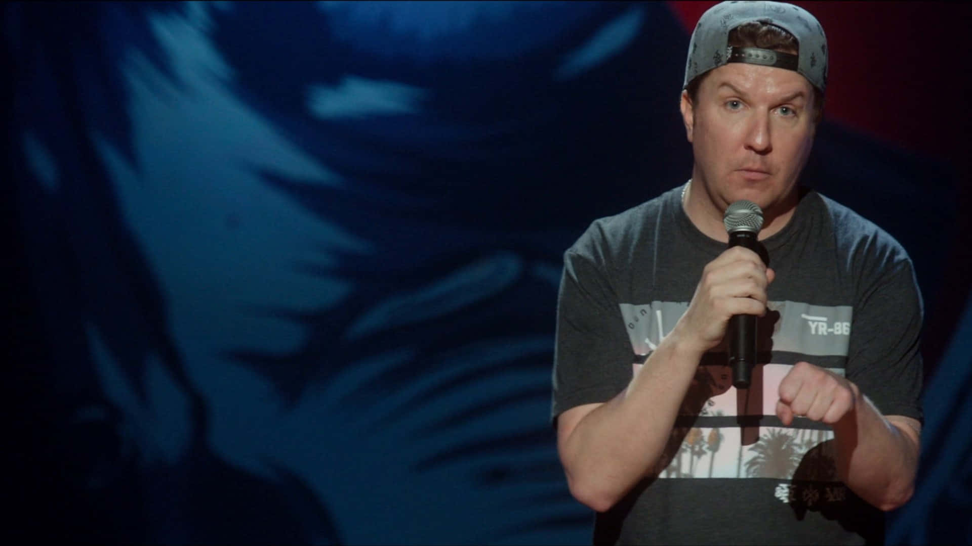 Nick Swardson 桌布 壁紙 — 一位戴帽子的喜劇演員在戲劇性的藍色燈光下，在模糊的抽象背景前，於舞台上握著麥克風。
