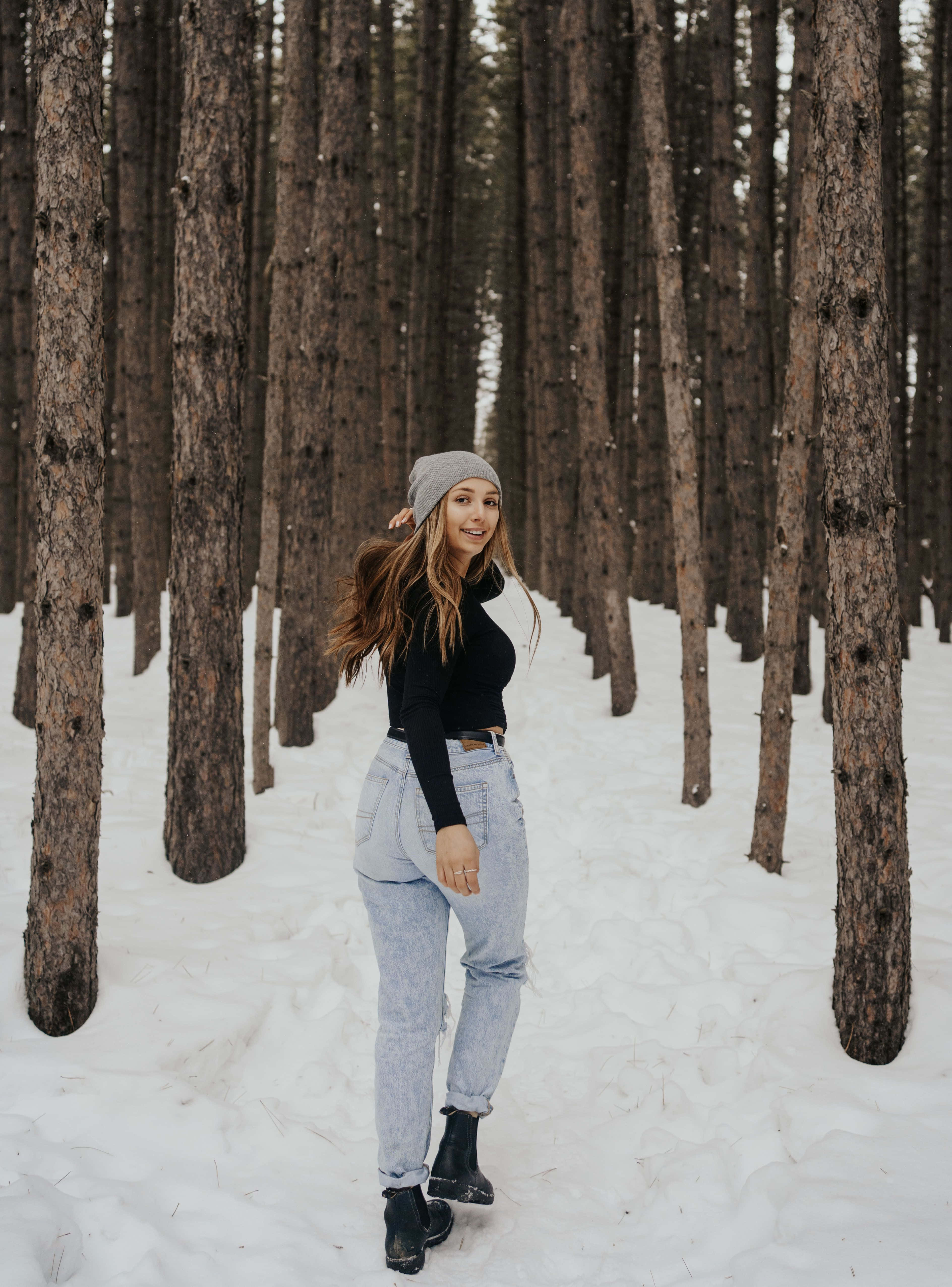 Winter Photography 桌布 壁紙 — 一位戴灰色冷帽、穿淺色牛仔褲的微笑女士站在被新雪覆蓋的高大松樹之間。