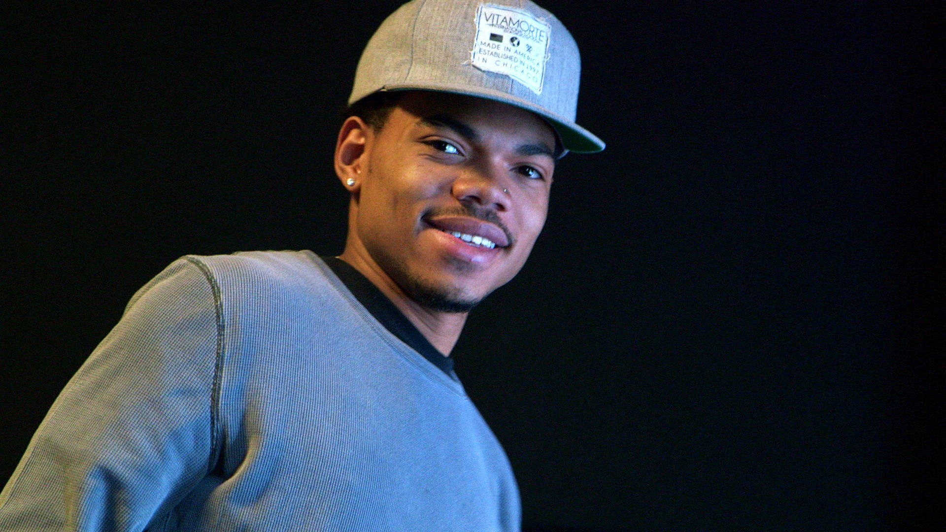 Chance the Rapper Vitamorte 壁紙 — 一位戴灰色鴨舌帽和穿毛衣的年輕男子在深色背景前自信地微笑。