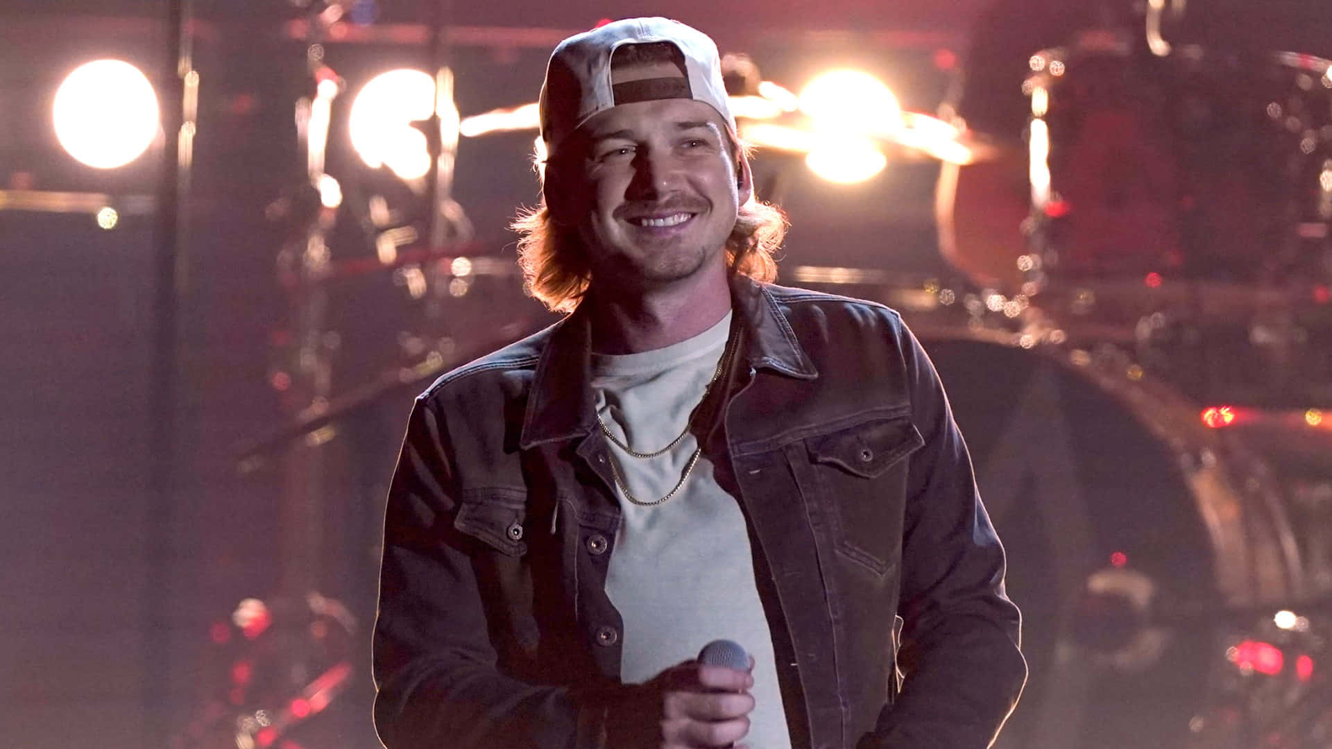Morgan Wallen 桌布 壁紙 — 一位戴白色鴨舌帽、身穿牛仔褸的男歌手微笑著，在溫暖燈光的舞台上，身後有鼓組和聚光燈，手持咪高峰。