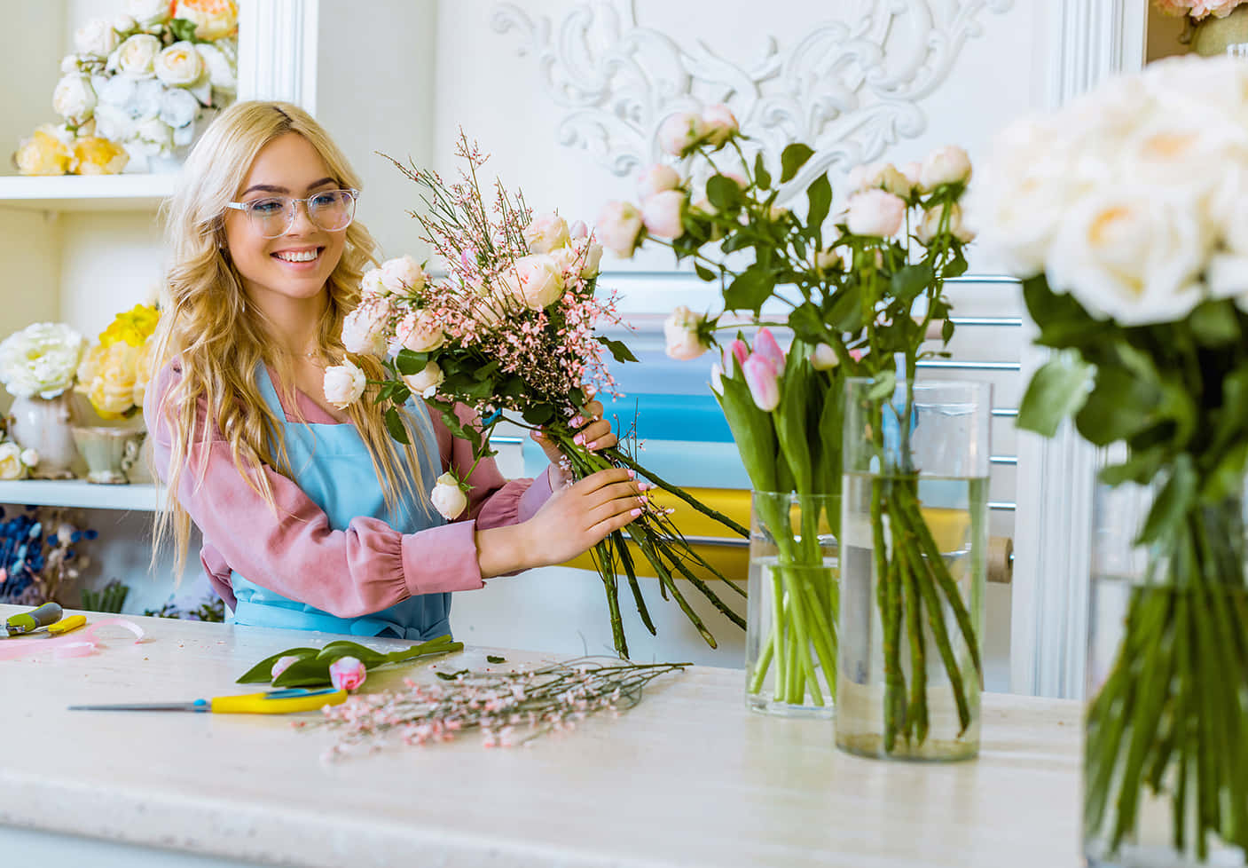 Florist 桌布 壁紙 — 一位戴眼鏡、微笑的金髮花店店員在明亮的鮮花店裡整理一束粉紅和白色的玫瑰。