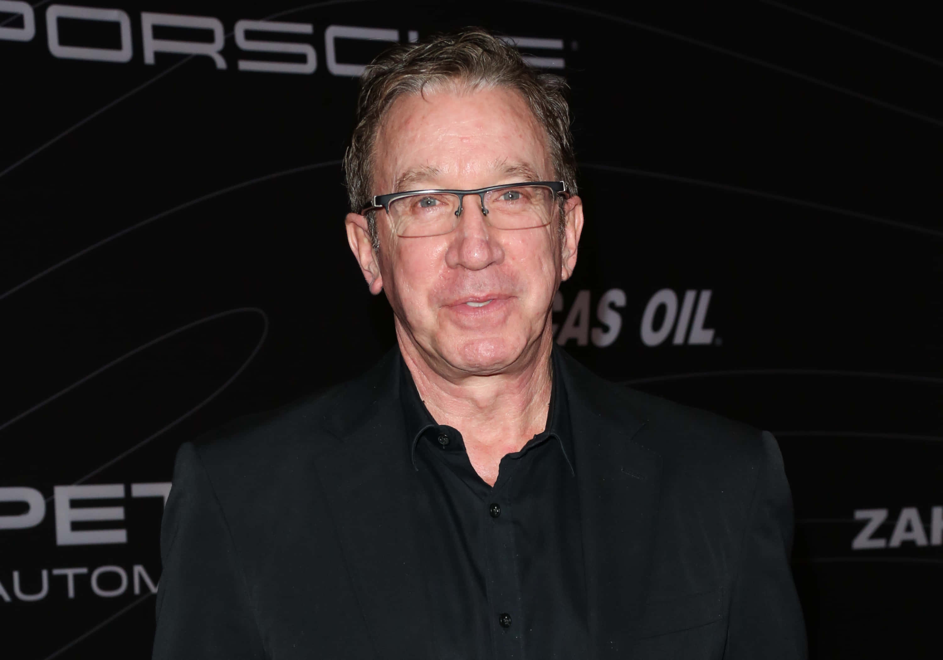 Tim Allen 桌布 壁紙 — 一位戴眼鏡、深色頭髮的中年男子在黑色保時捷品牌背景前對著鏡頭微笑。