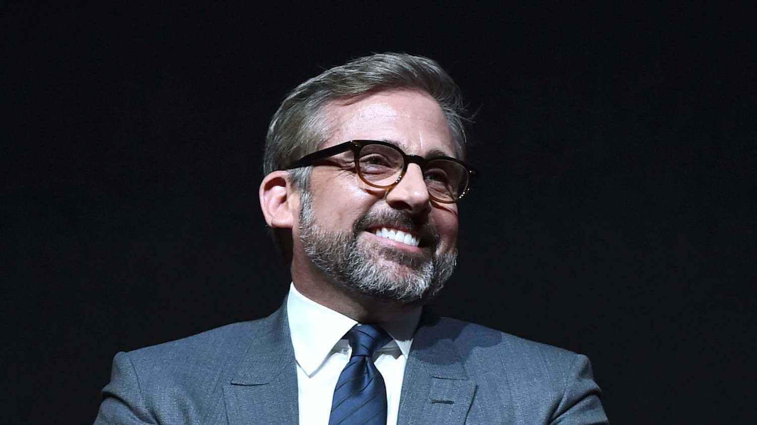 steve carell 桌布 壁紙 — 一位戴眼鏡、留鬍鬚的男士，身穿深色西裝，在黑色背景前溫暖地微笑。