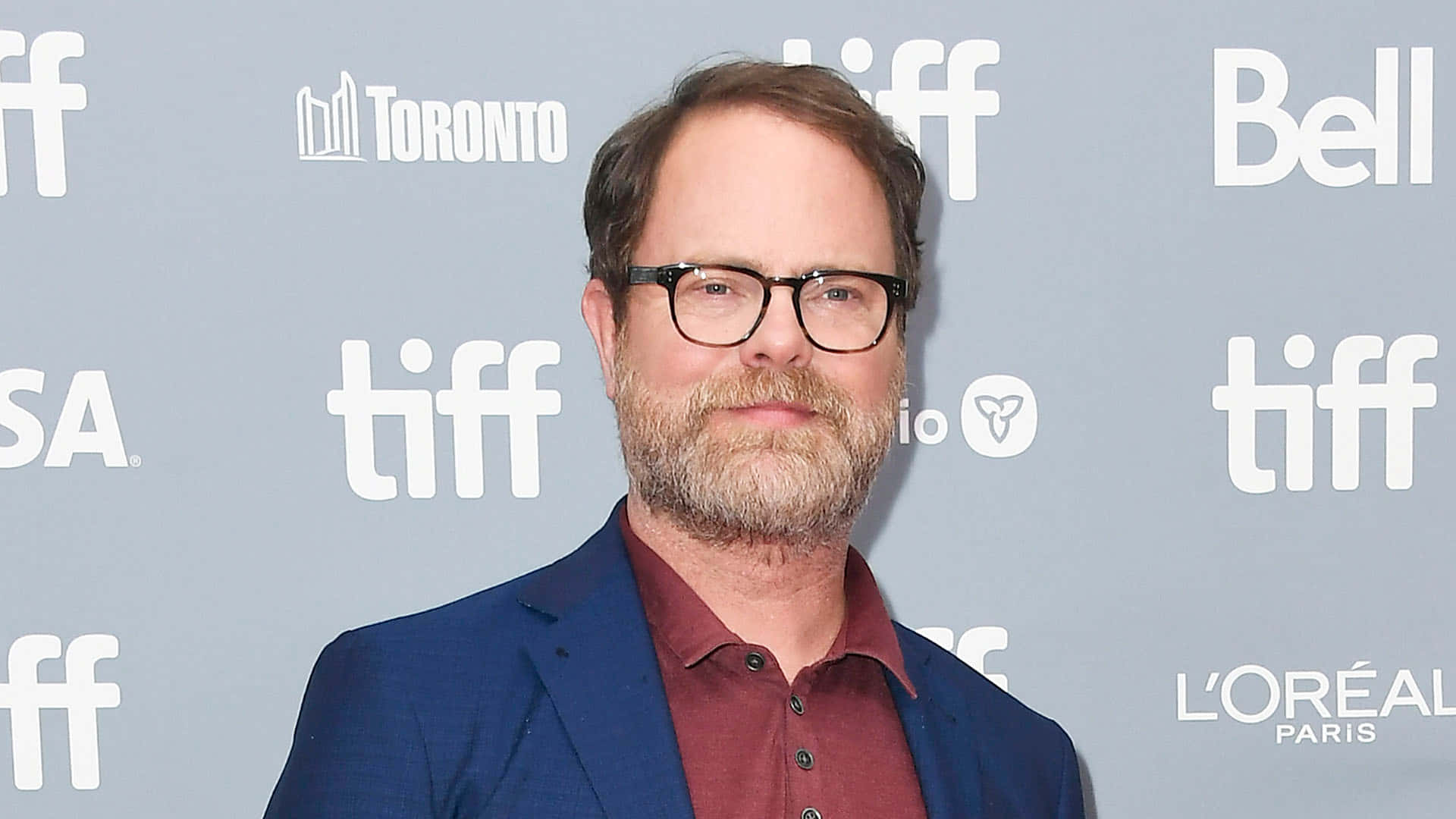 rainn wilson 桌布 壁紙 — 一位戴眼鏡、留鬍鬚的男子，身穿藍色西裝外套和栗色襯衫，背景為灰色 TIFF。