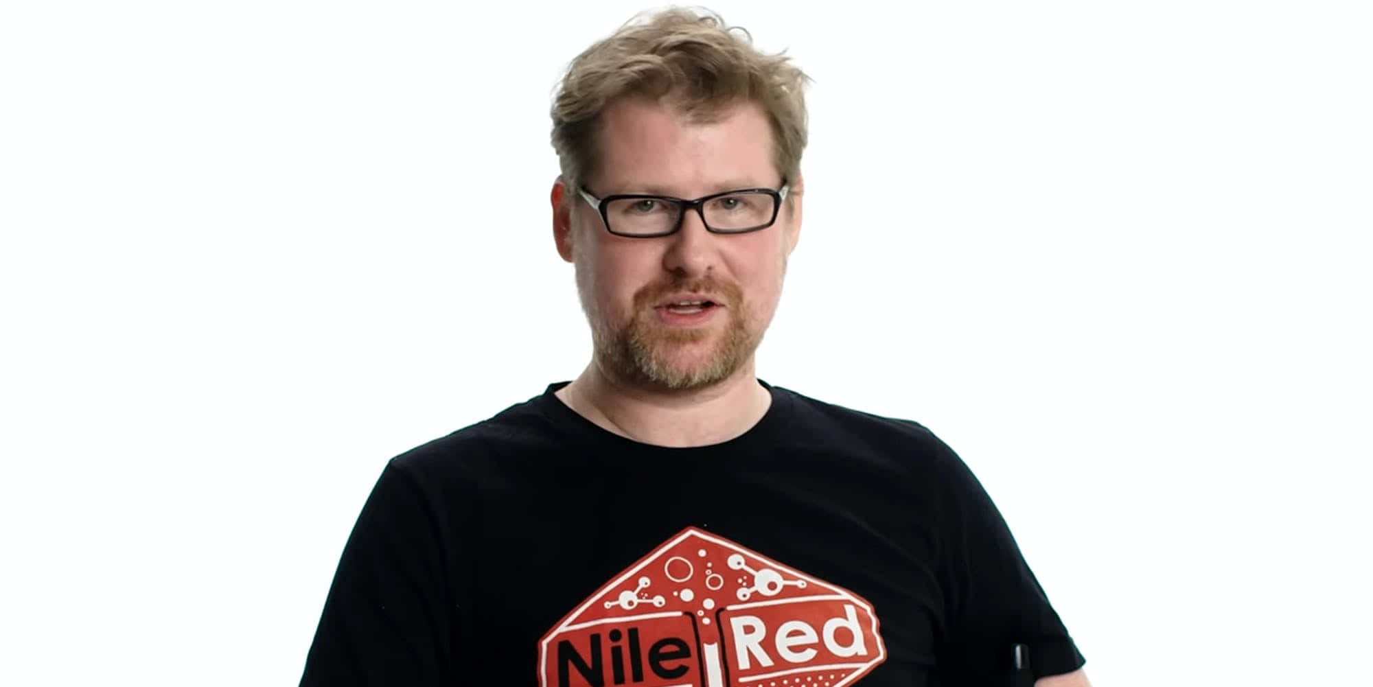 Justin Roiland 桌布 壁紙 — 一位戴眼鏡、留鬍鬚的男子，身穿黑色T恤，上面有紅色 'Nile Red' 標誌，背景為純白色。
