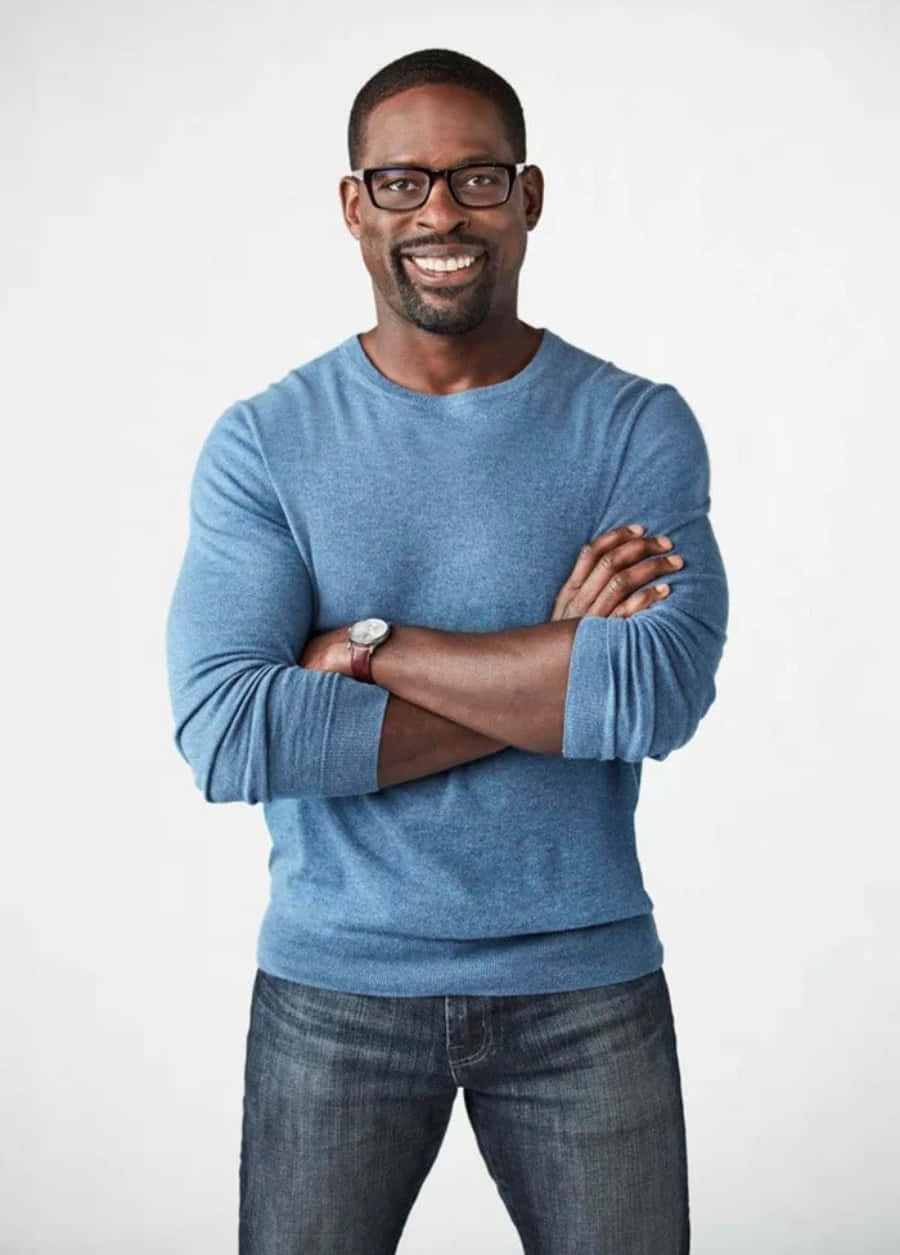 Sterling K Brown 桌布 壁紙 — 一位戴眼鏡、穿藍色毛衣的男士微笑著，自信地交叉雙臂站在純白色背景前。