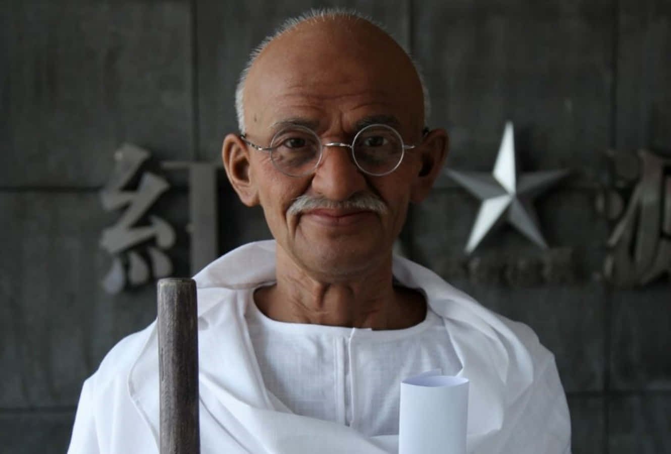 Mahatma Gandhi 桌布 壁紙 — 一位戴眼鏡、身穿白色多蒂的長者，在帶有金屬星星和漢字符號的深色牆壁前，手持杖和卷軸。