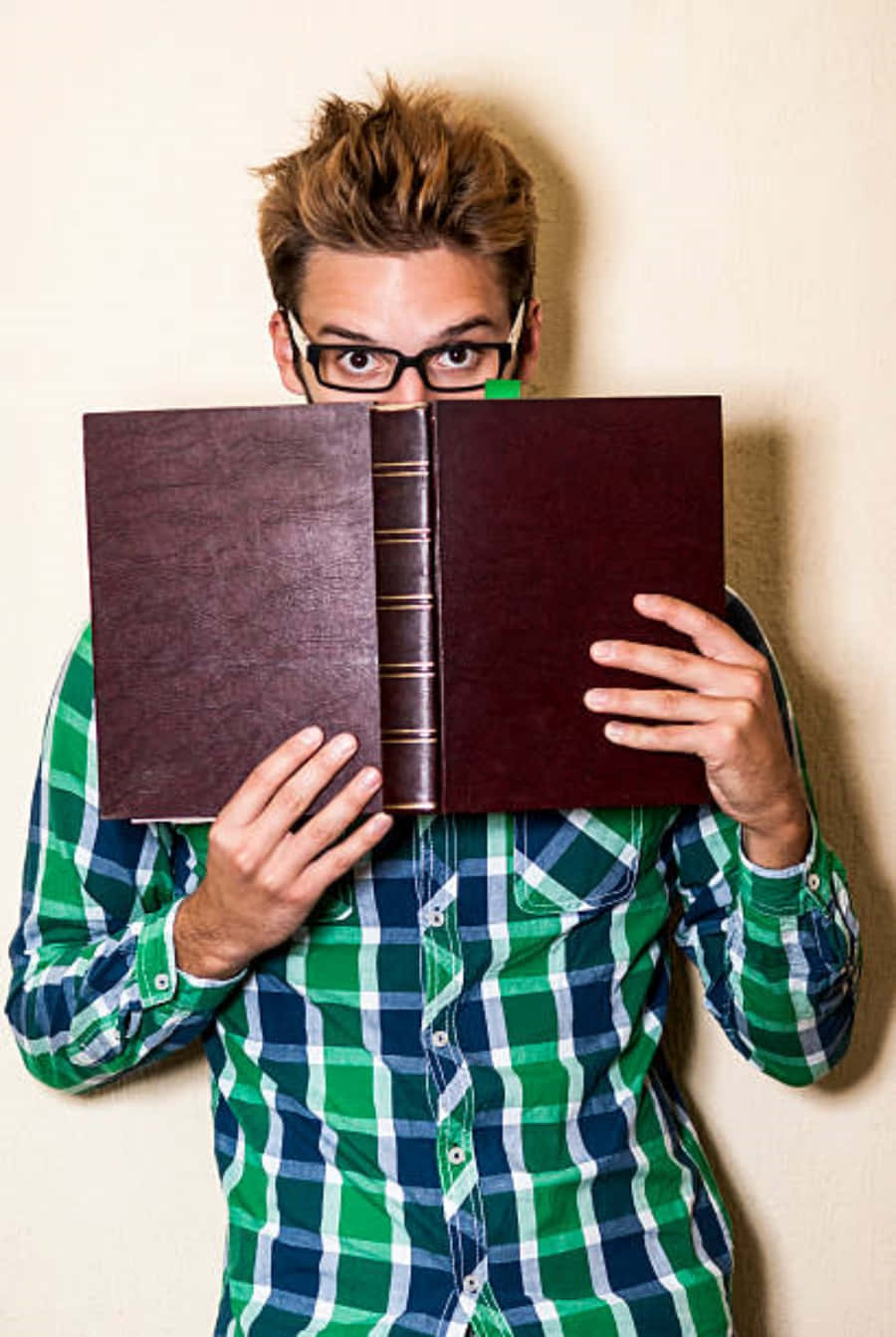 書呆子 桌布 壁紙 — 一位戴眼鏡、身穿綠色格仔恤衫的年輕男子，將臉藏在一本打開的大書後面，同時頑皮地看著鏡頭。
