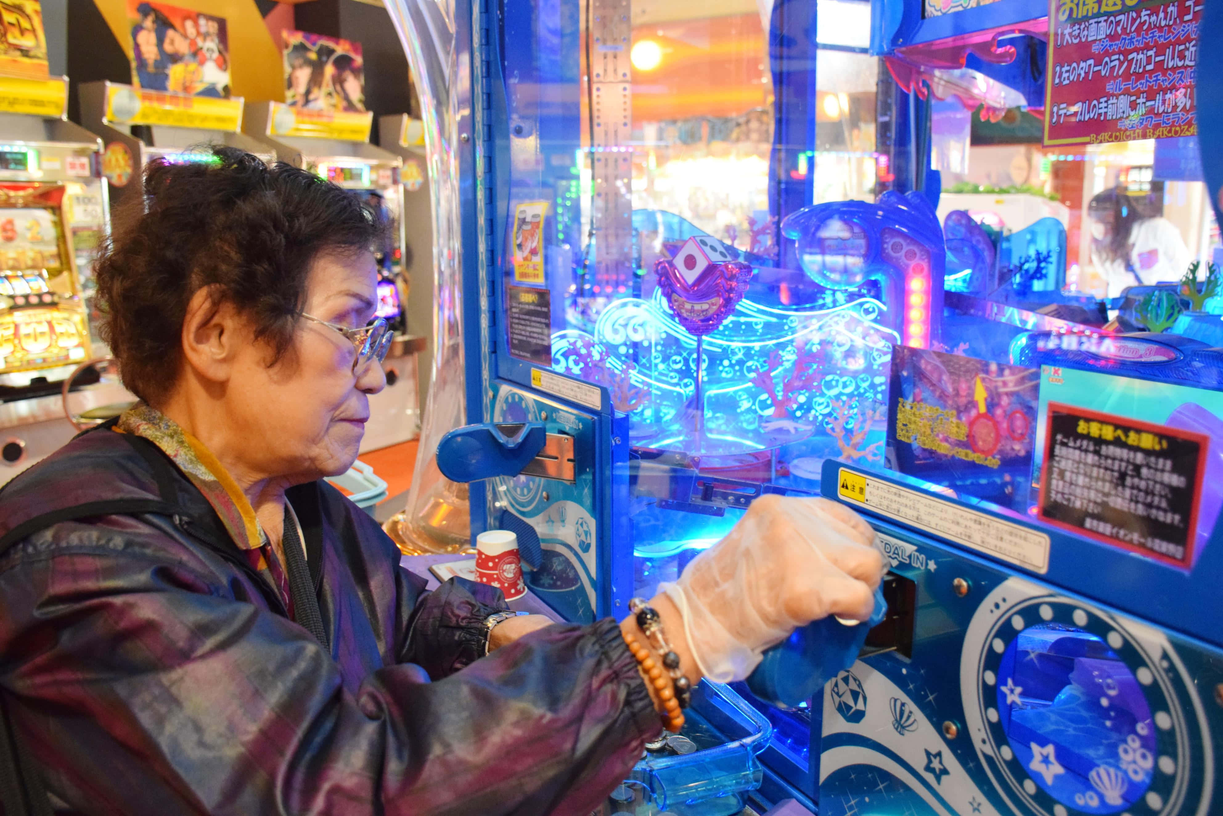 Arcade Games 桌布 壁紙 — 一位戴眼鏡和膠手套的老年女士，在熱鬧的遊戲機中心專注地玩著發光的藍色夾公仔機。