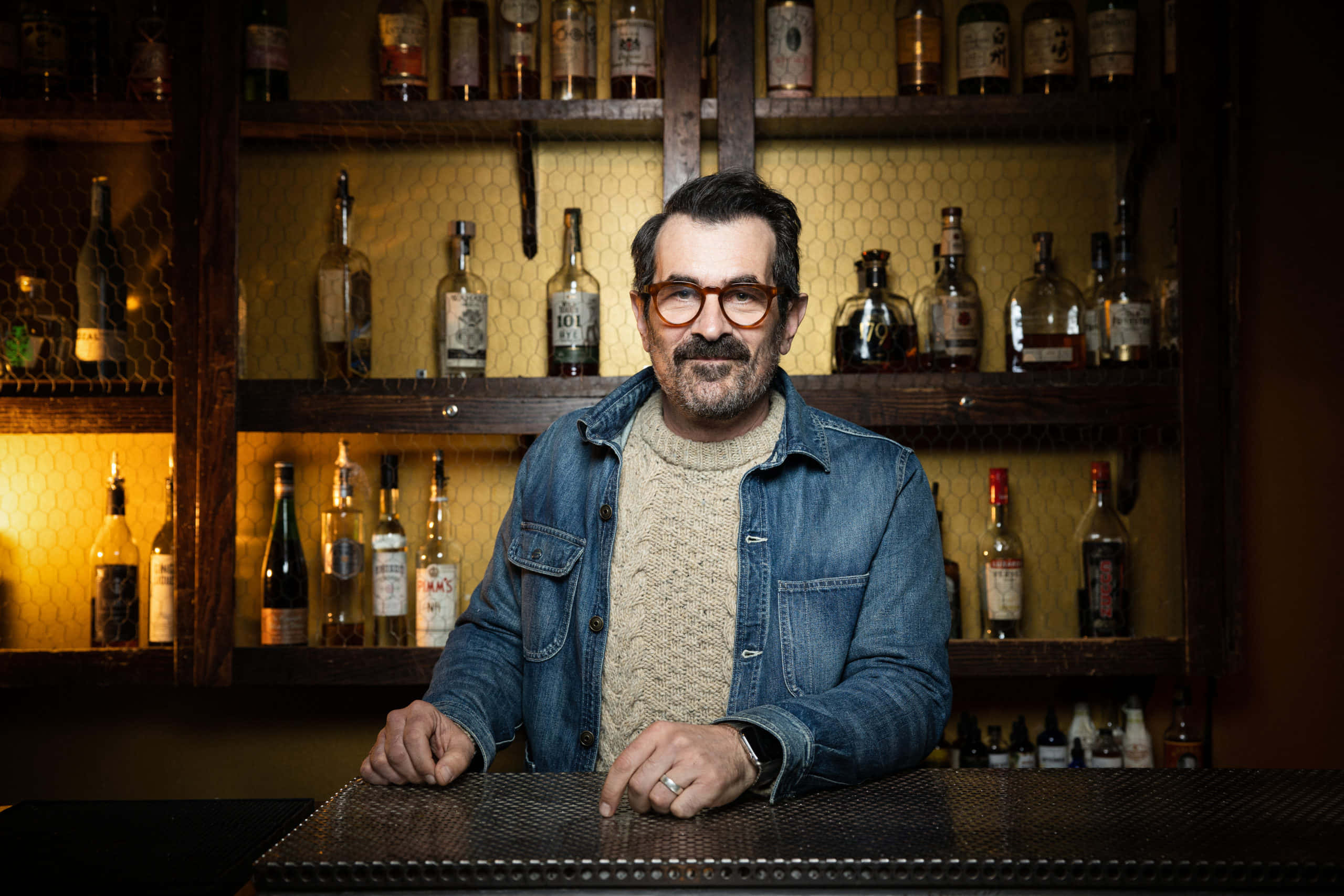 Ty Burrell 桌布 壁紙 — 一位戴眼鏡留鬍鬚嘅男人坐喺吧枱前，身後有暖色燈光照射嘅酒瓶架。